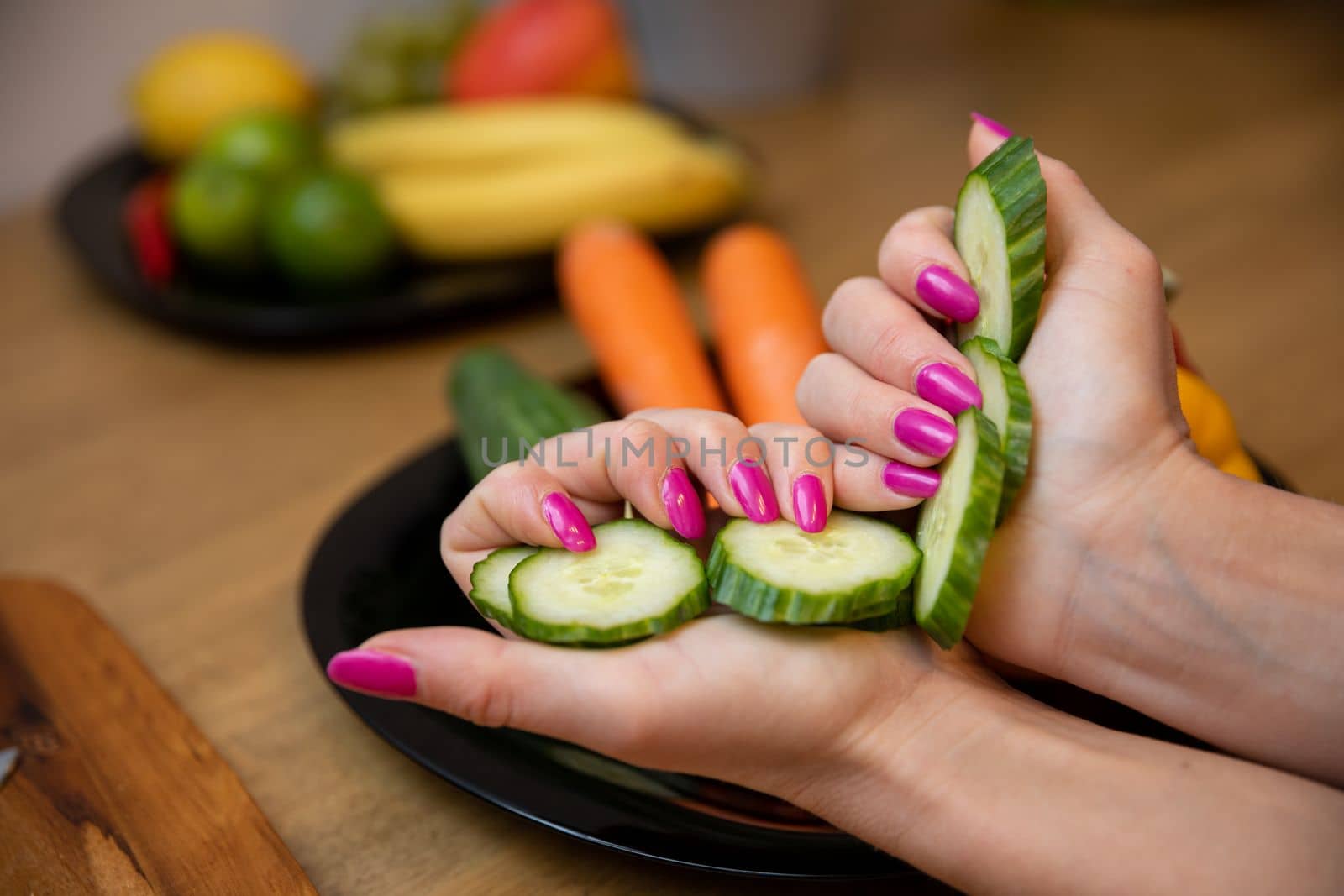 Close-up on female hands in foreground holding sliced cucumber. Blurred background in the background of plates with vegetables and fruits. The woman has her nails painted pink.