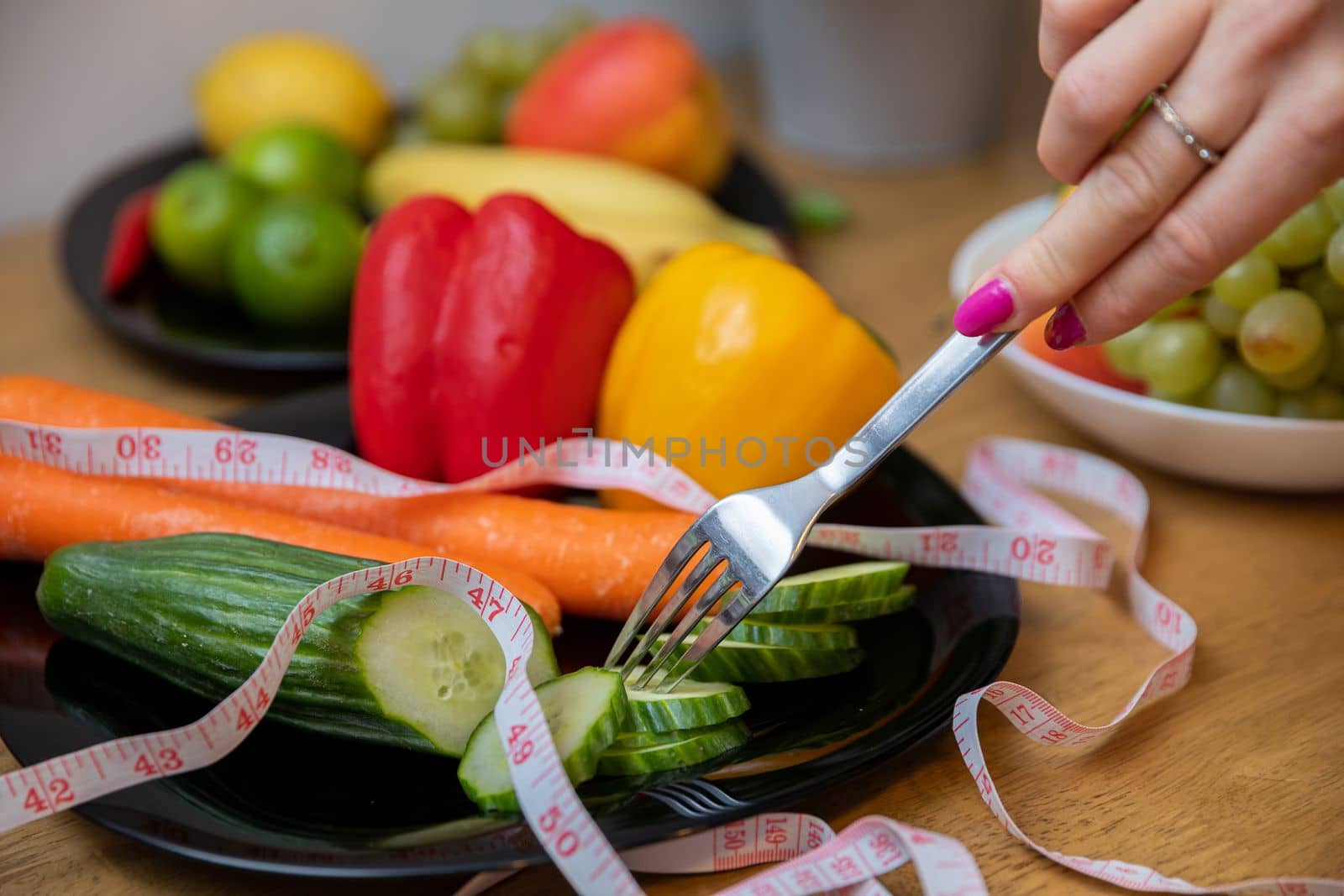 A plate with sliced cucumber and other vegetables, visible up close. A measure tape lies on top of the vegetables. A woman's hand holding a fork brings it closer to the vegetables.