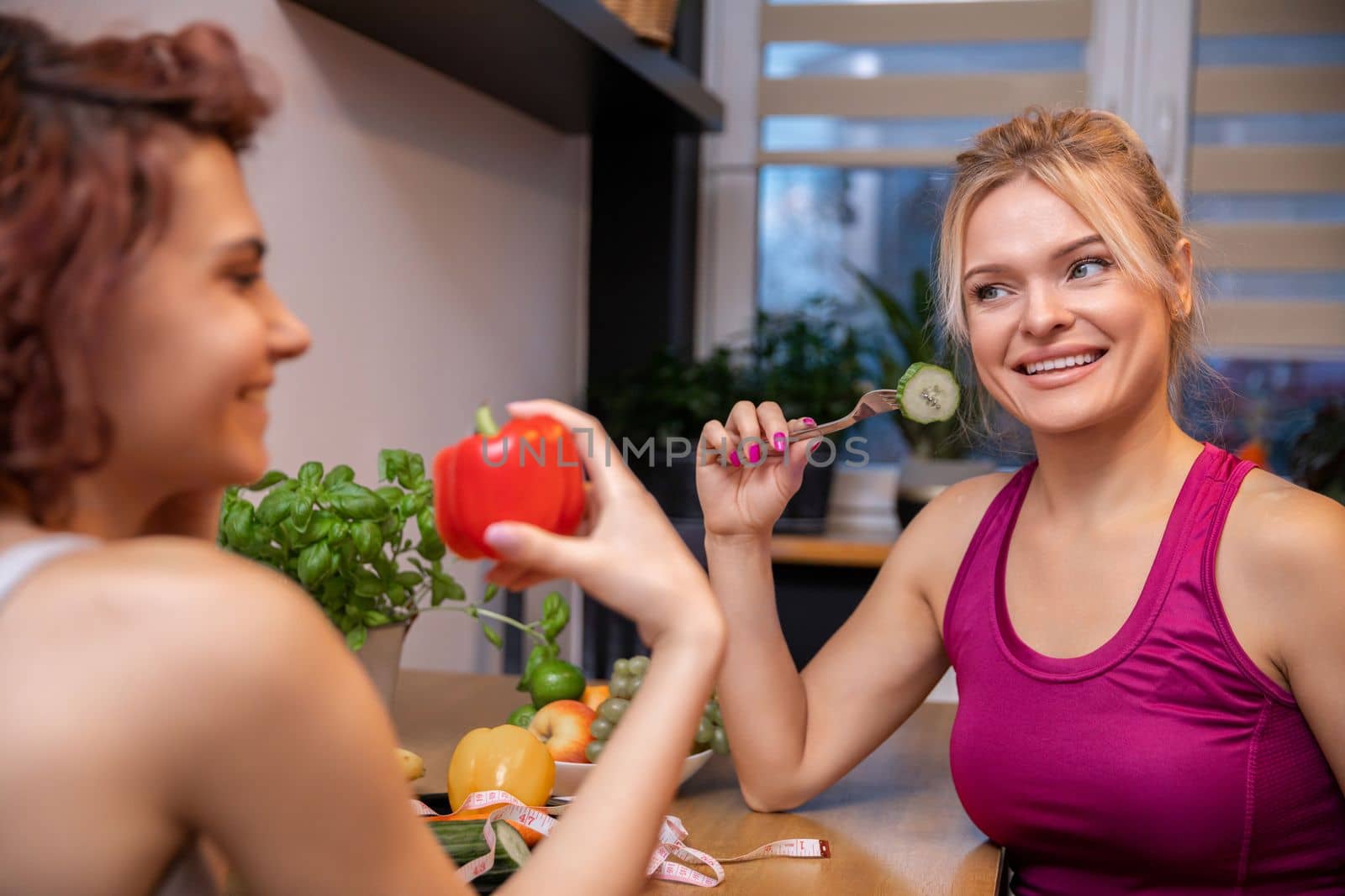 Two women are sitting in the kitchen at a table facing each other. The woman in the foreground holds a red bell pepper, the woman in the background holds a fork with a cucumber slice.