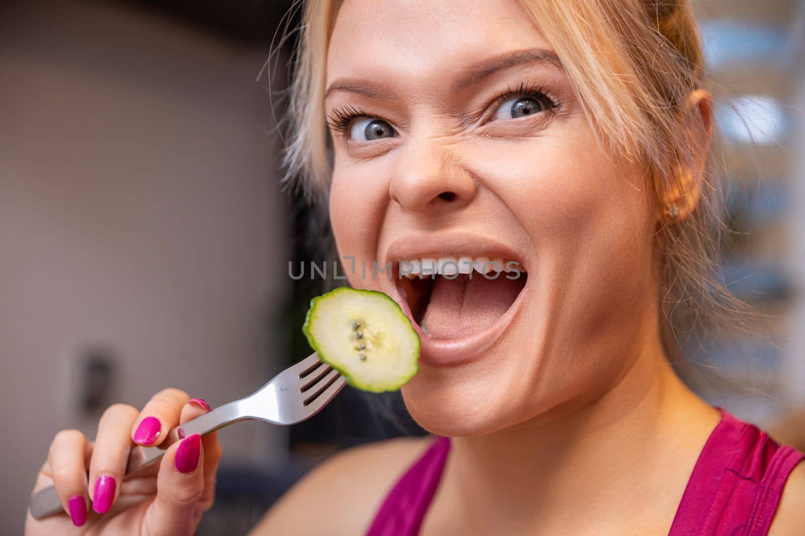 Large close-up on the face of a blonde woman with her mouth open. The woman holds a fork with a scooped cucumber slice with skin and brings it close to her open mouth.
