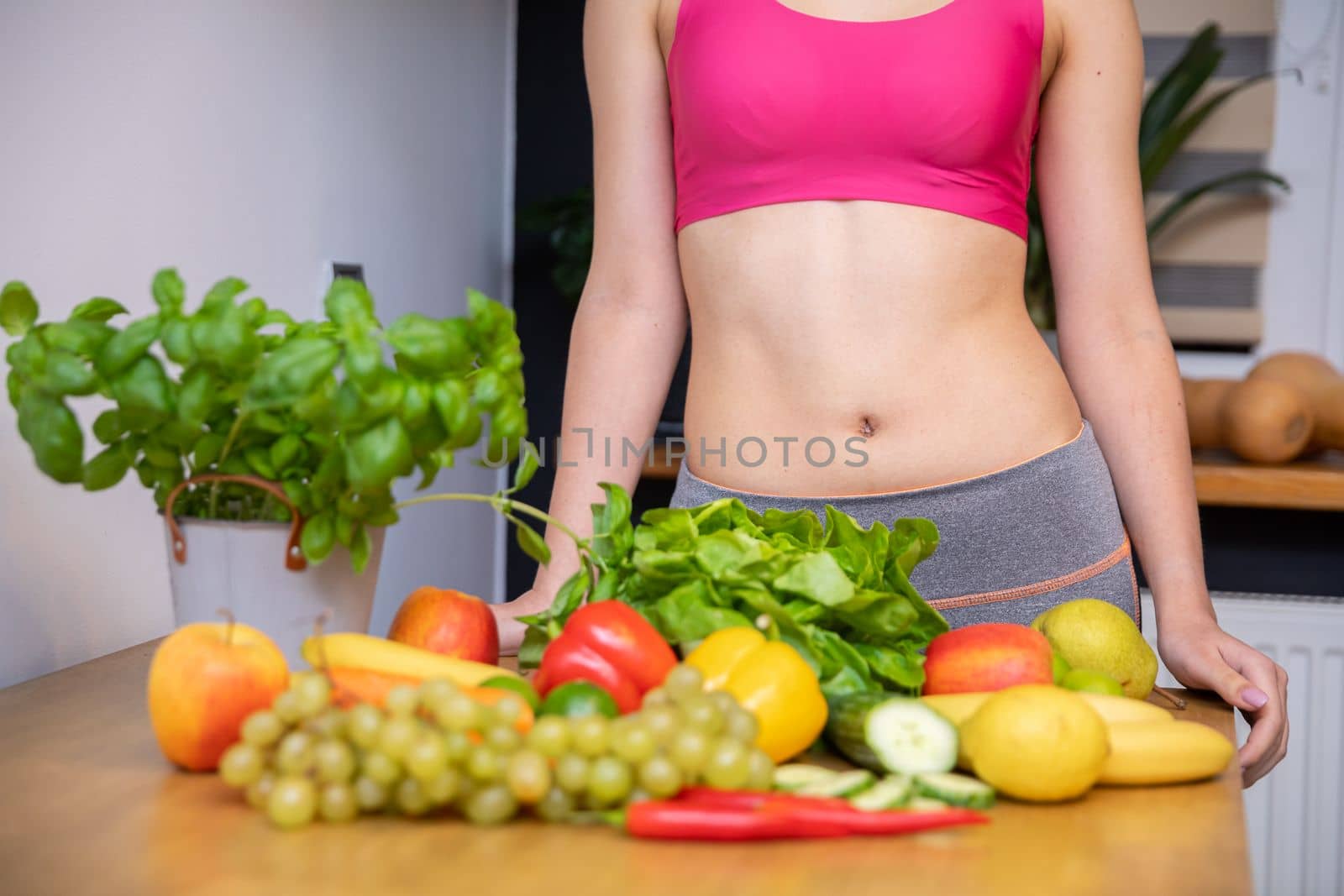 Lots of vegetables and fruits lying on the kitchen table. Behind the table a silhouette of a woman with an exposed belly in a tight pink sports top leaning against the table top