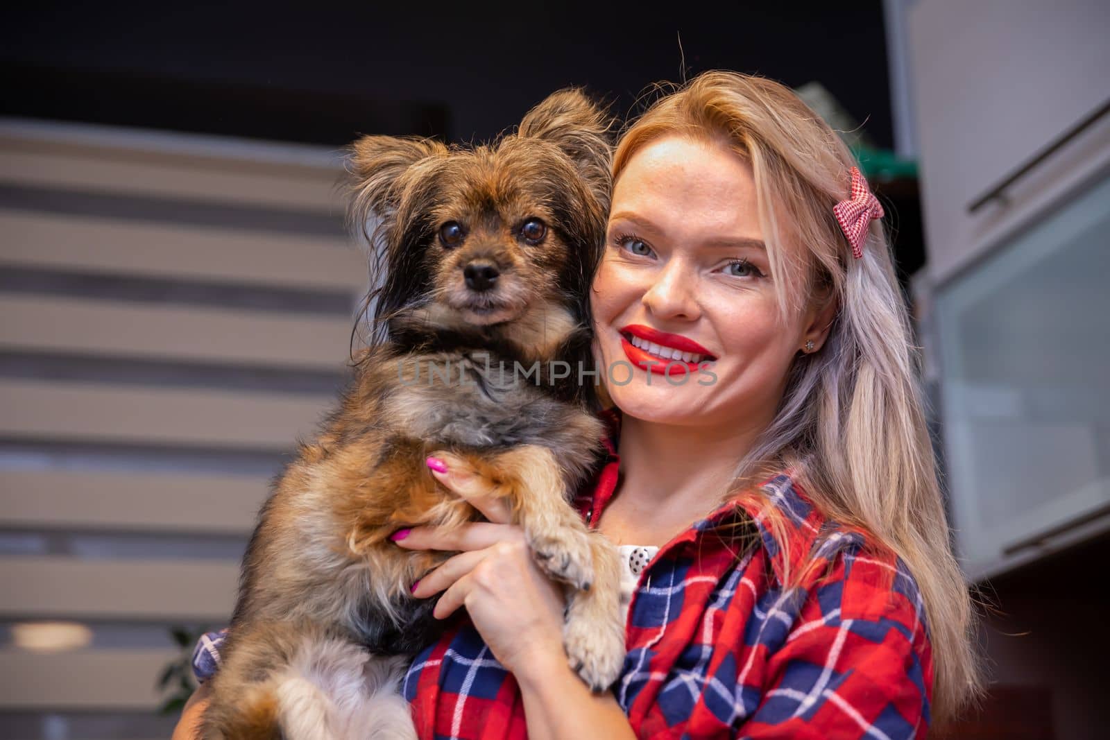 A small brown shaggy dog sitting in the arms of its owner. A woman with red lipstick cuddling a fluffy dog in her arms. A mutt from a shelter held in her arms