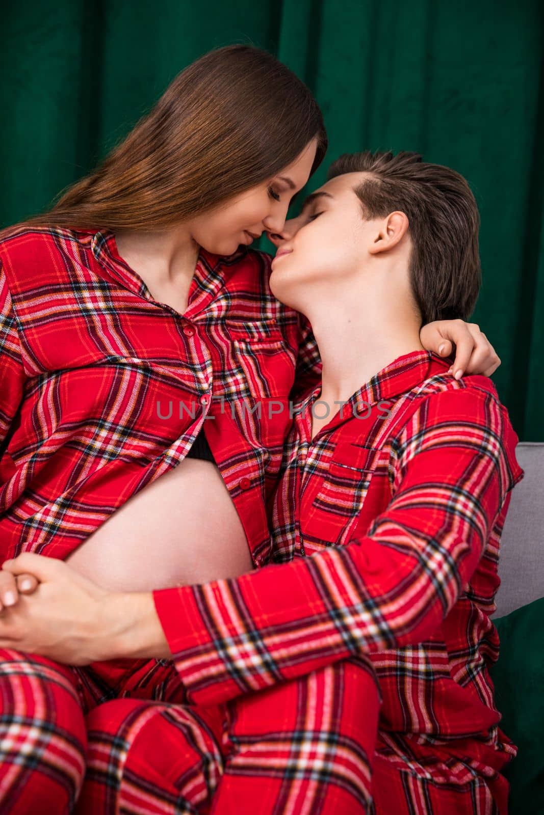 Man and woman in love turn their faces towards each other aiming for a kiss. Girl sitting on boyfriend's lap, revealing pregnant belly. The couple wears the same red checkered pajamas.