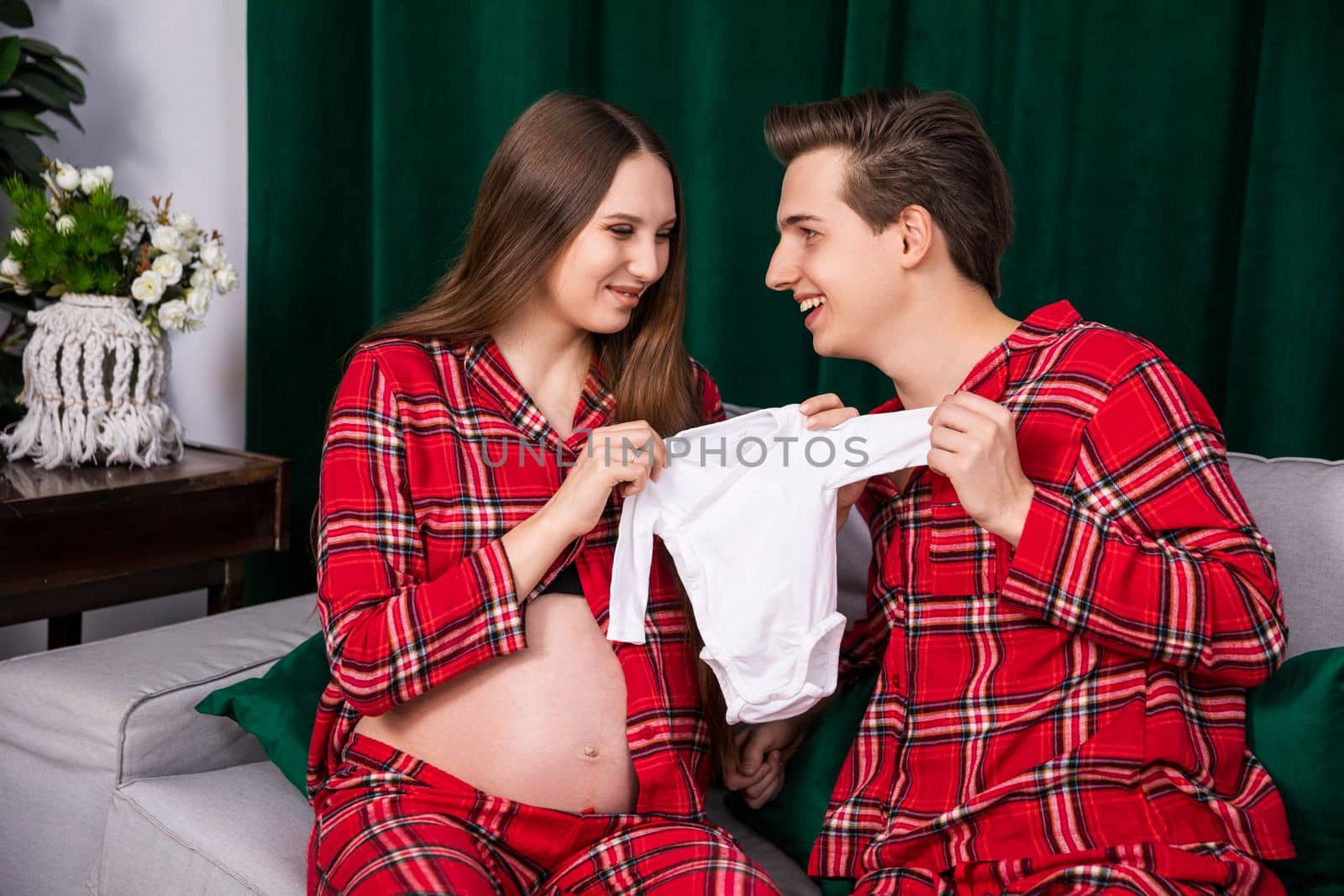 A pregnant woman with an exposed belly sits next to her man and together they hold a baby's light-colored sleepwear.