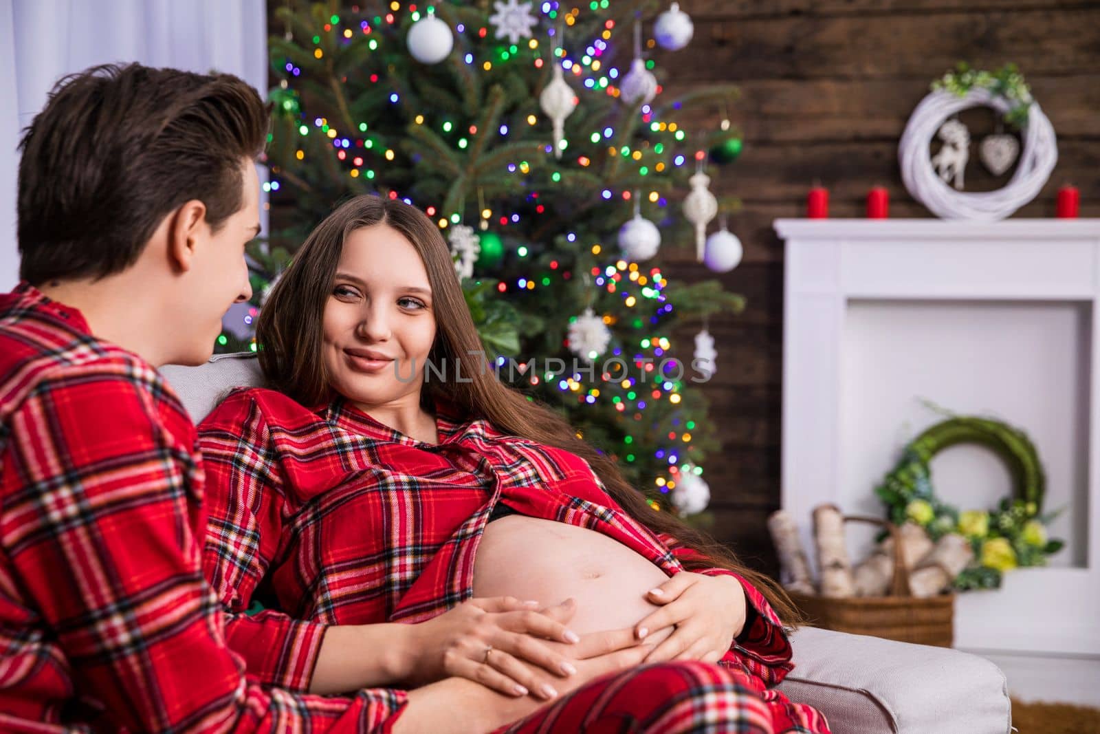 A pregnant woman sits next to her husband and smiles. In the background is a blurry dressed Christmas tree. The mother-to-be keeps her hands on her exposed pregnant belly.