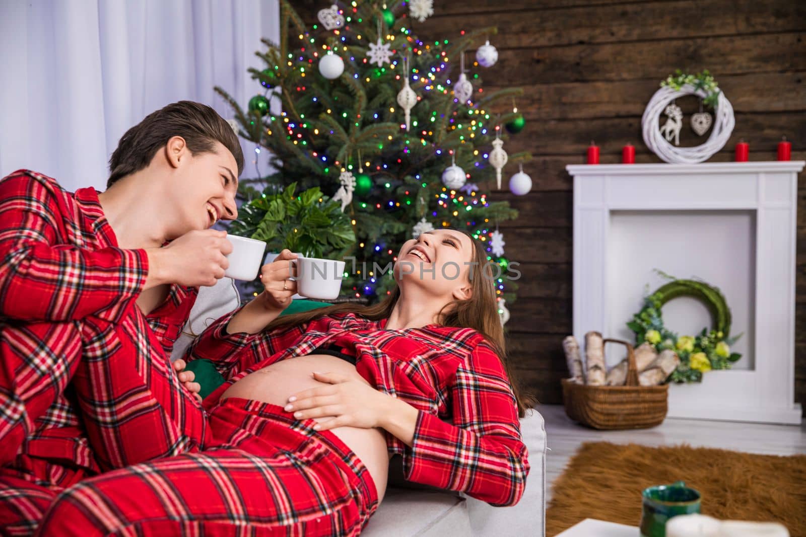 The happy parents-to-be are lying on the couch and laughing while holding white teacups, against a backdrop of a dressed Christmas tree with colorful lights. The couple is wearing the same checkered pajamas.