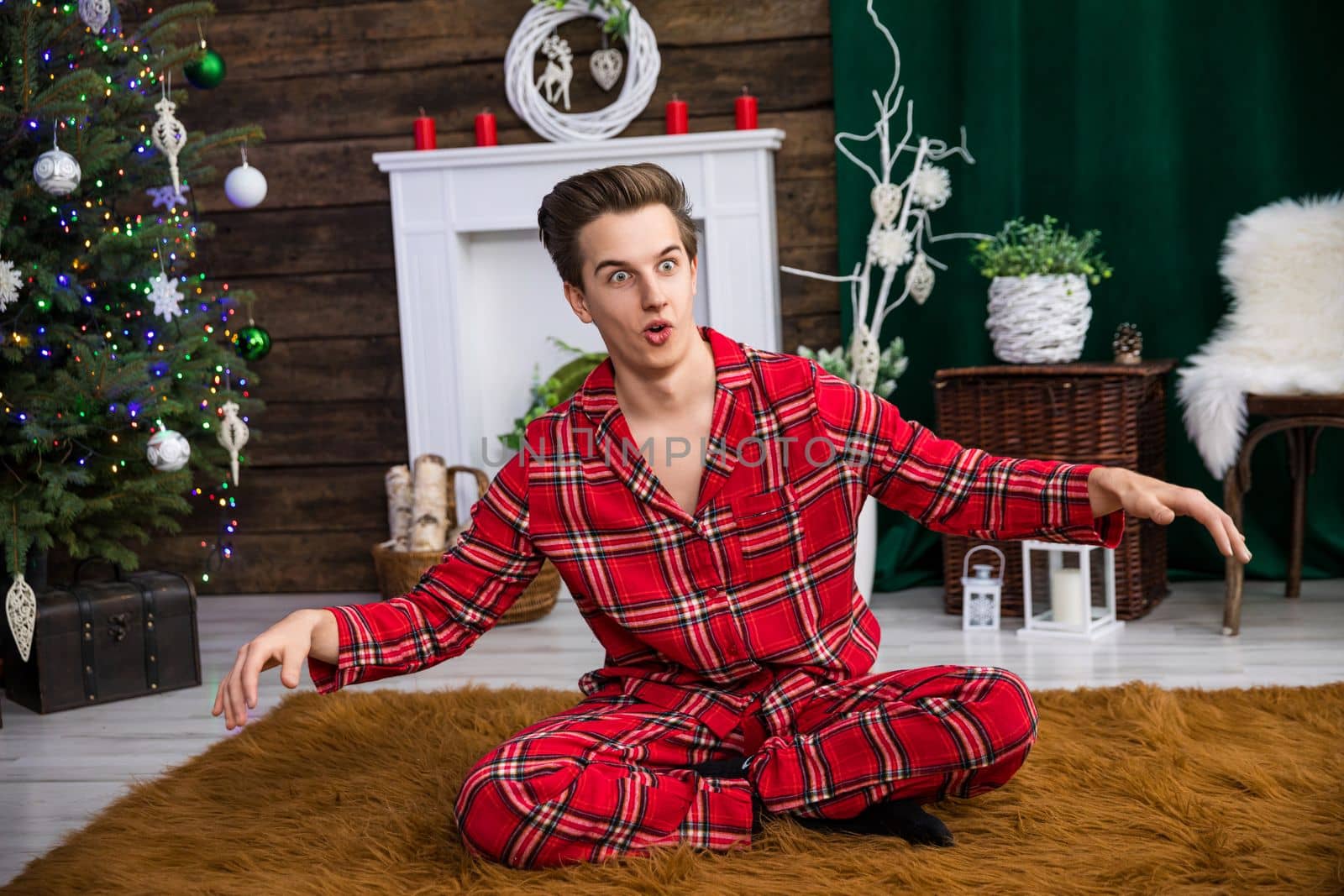 A man sits with his legs crossed on a brown carpet with long hair. He is prancing around and making funny faces, his hands raised. In the background a dressed Christmas tree and other holiday decorations