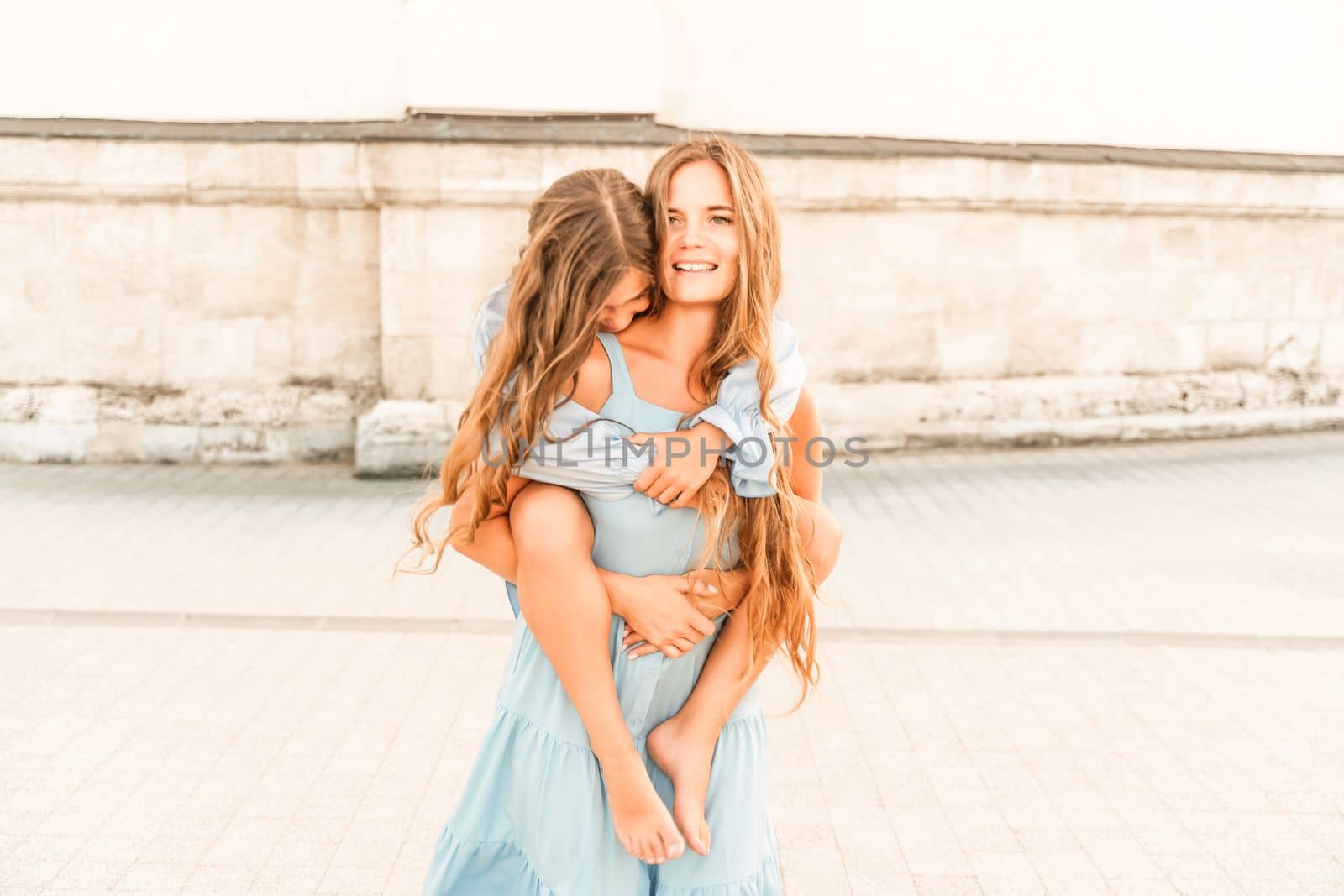 Mother of the daughter walks playing. Mother holds the girl on her back, holding her legs, and her daughter hugs her by the shoulders. Dressed in blue dresses