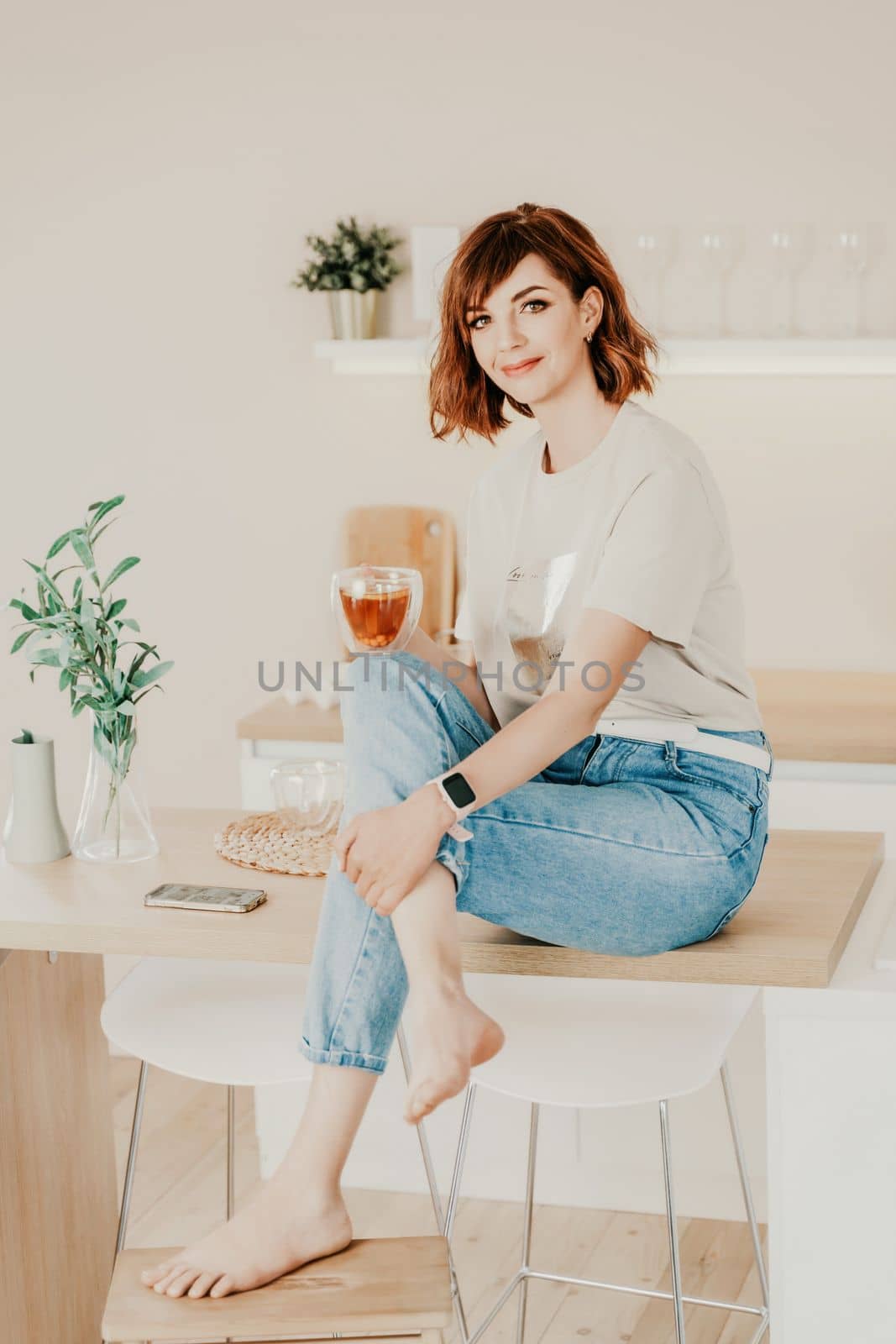 Woman kitchen tea. Carefree woman rests in a cozy kitchen, drinks tea, sits on the table and dreams, free space. Woman holding cup while enjoying hot drink at home.