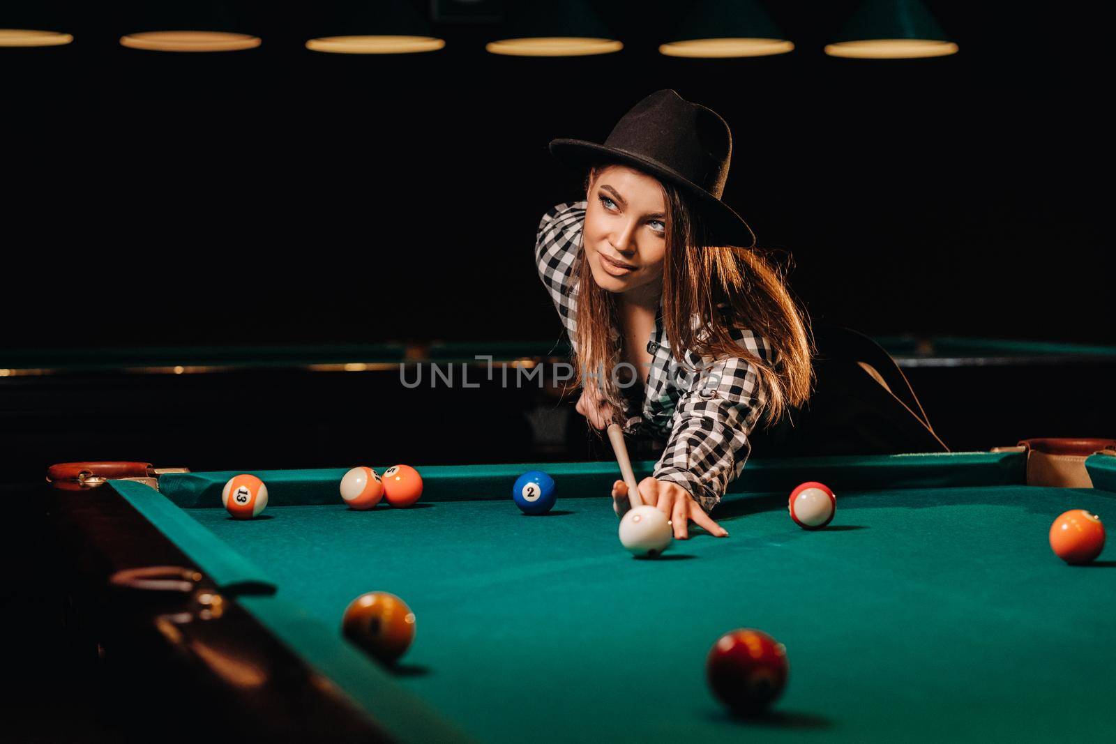 A girl in a hat in a billiard club with a cue in her hands hits a ball.Playing billiards by Lobachad
