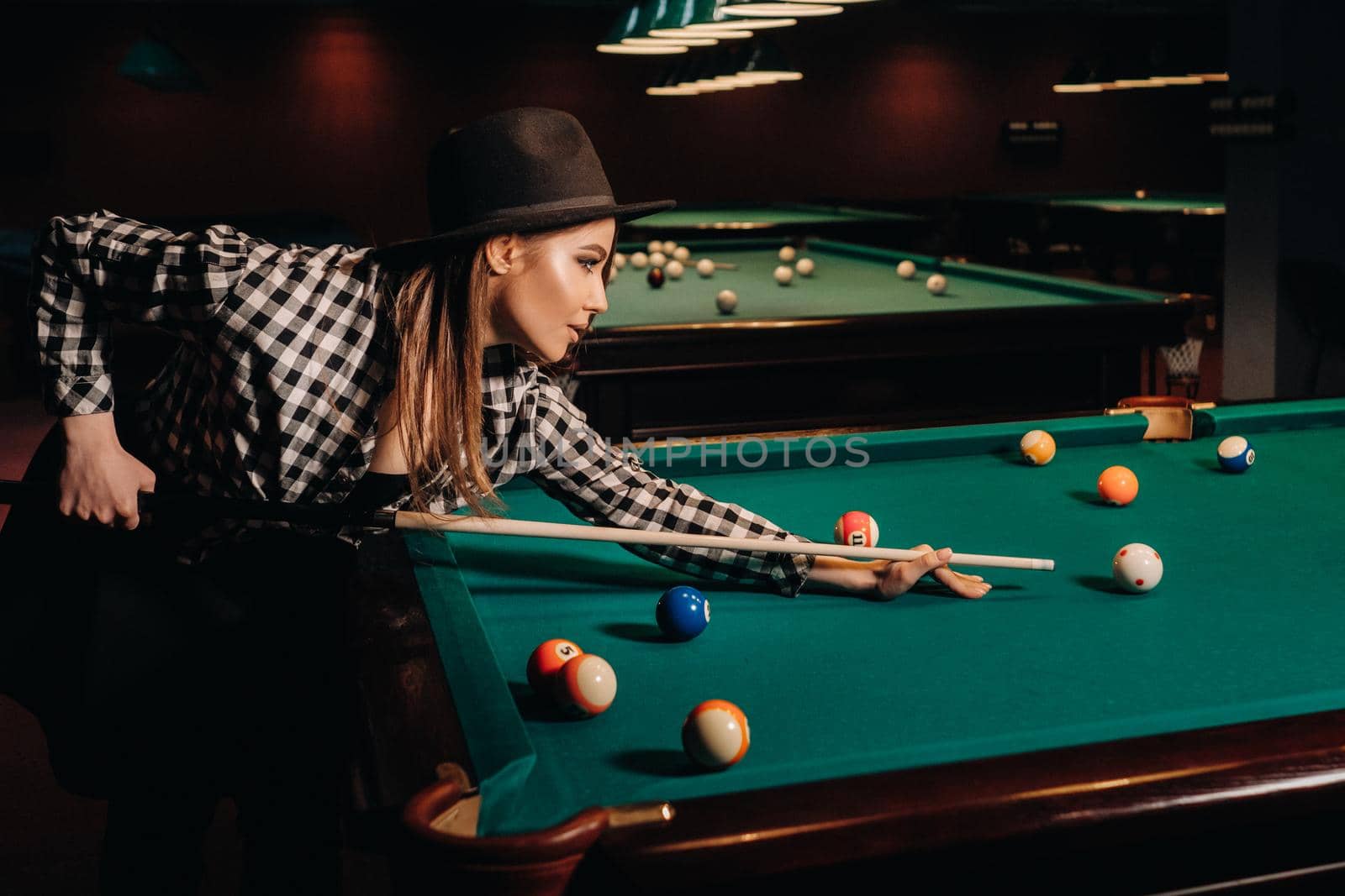 A girl in a hat in a billiard club with a cue in her hands hits a ball.Playing pool by Lobachad