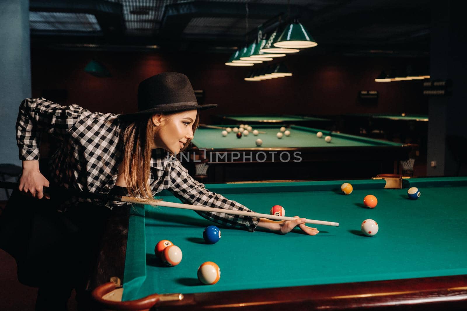 A girl in a hat in a billiard club with a cue in her hands hits a ball.Playing pool.