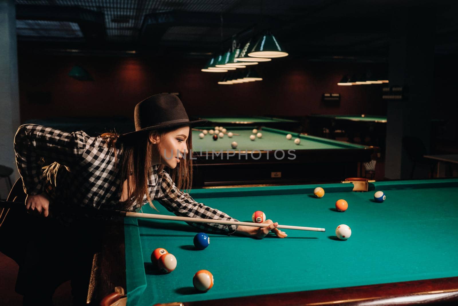 A girl in a hat in a billiard club with a cue in her hands hits a ball.Playing pool by Lobachad
