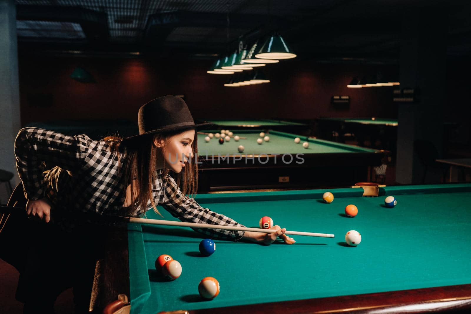 A girl in a hat in a billiard club with a cue in her hands hits a ball.Playing pool.