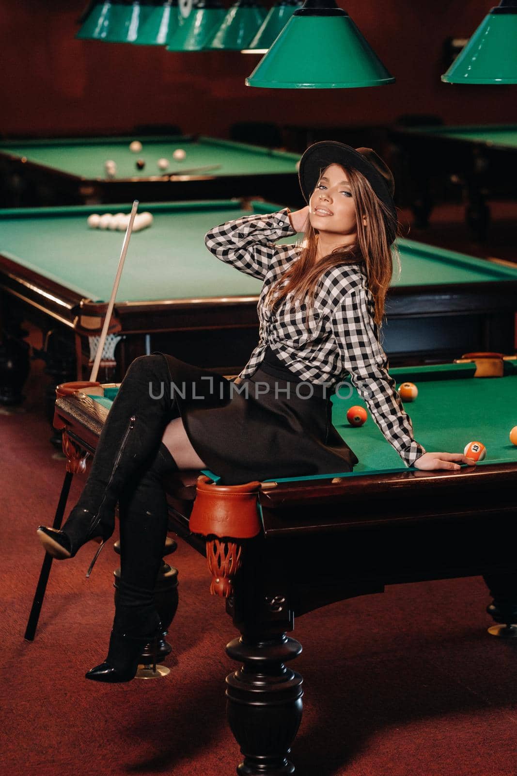 A girl in a hat in a billiard club sits on a billiard table.Playing pool by Lobachad