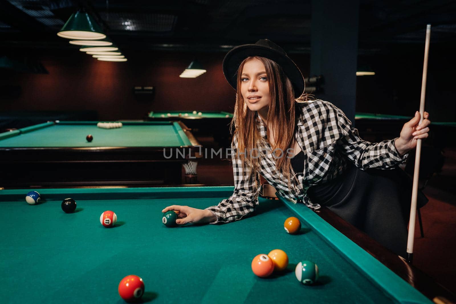 A girl in a hat in a billiard club with a cue and balls in her hands.Playing pool.
