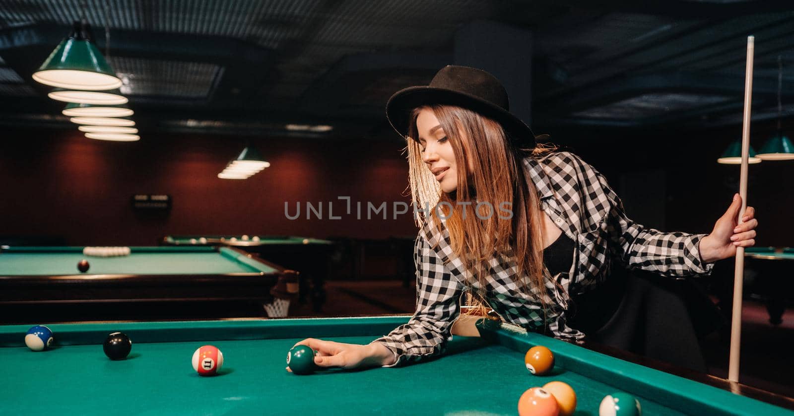 A girl in a hat in a billiard club with a cue and balls in her hands.Playing pool.