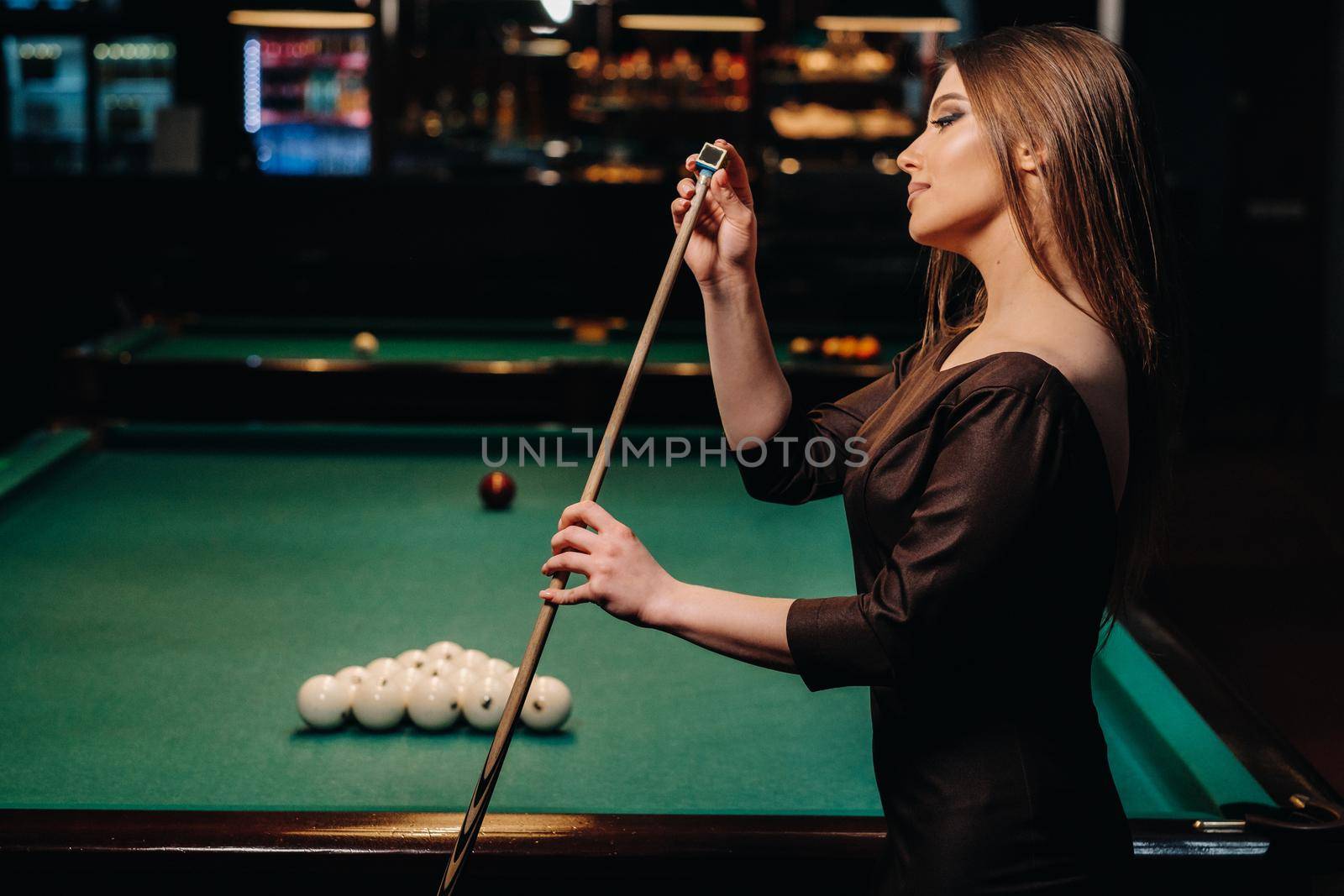 A girl in a dress stands with a cue in her hands and cleans it with chalk in a billiard club with balls in her hands.Playing billiards by Lobachad