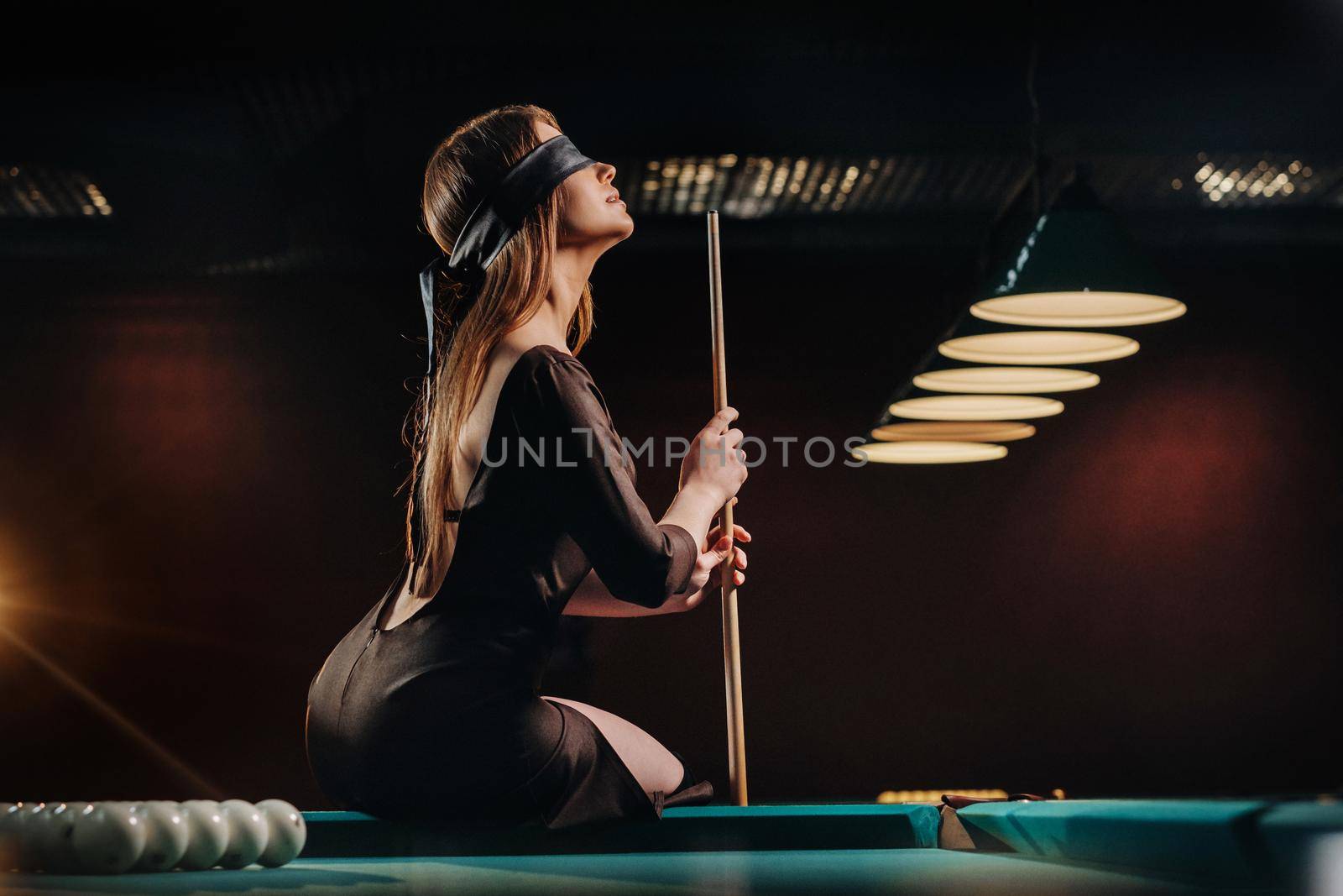 A girl with a blindfold and a cue in her hands is sitting on a table in a billiard club.Russian billiards by Lobachad