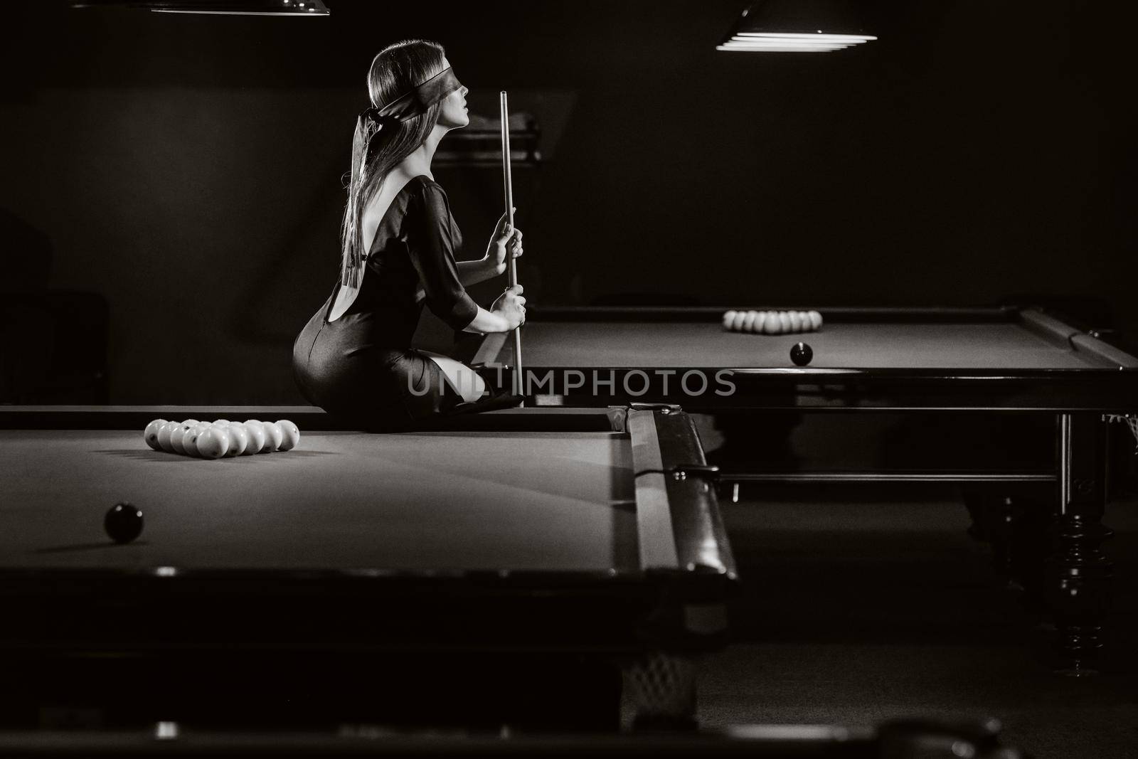 A girl with a blindfold and a cue in her hands is sitting on a table in a billiard club.Russian billiards. black and white photo.