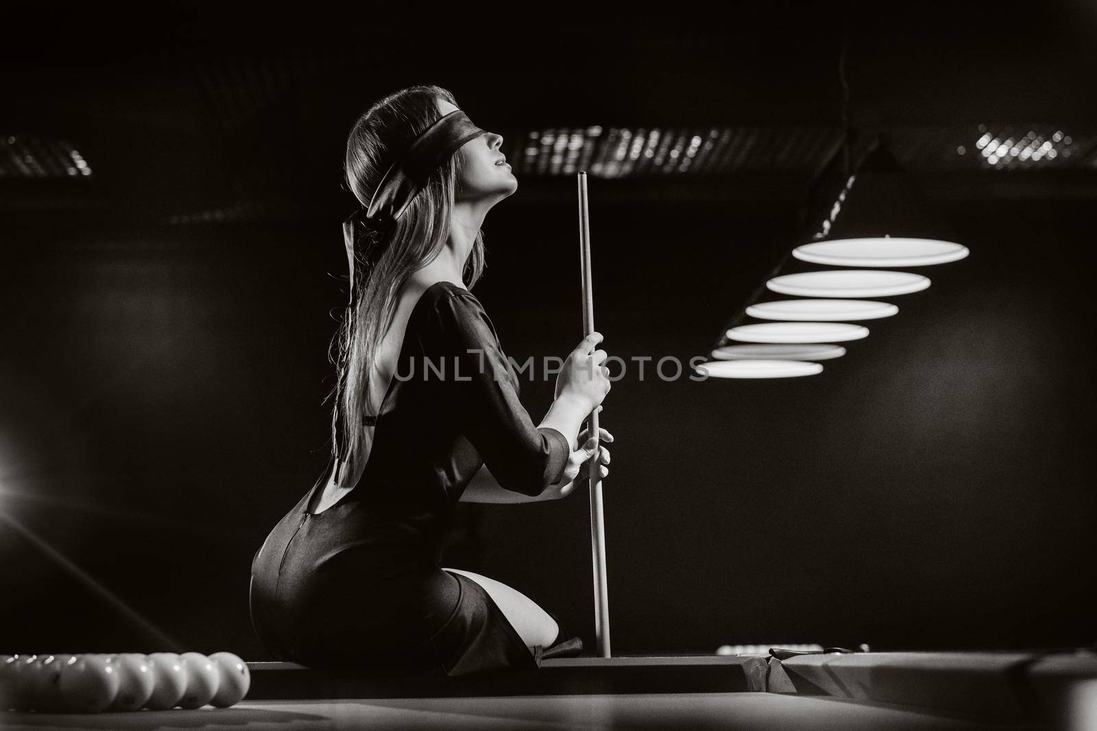 A girl with a blindfold and a cue in her hands is sitting on a table in a billiard club.Russian billiards. black and white photo by Lobachad