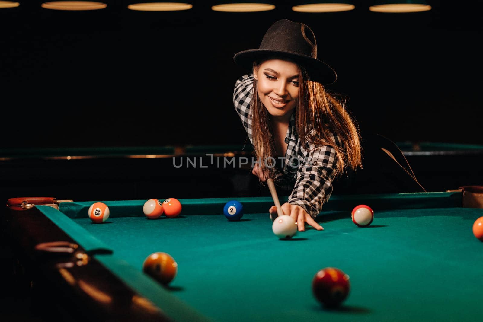 A girl in a hat in a billiard club with a cue in her hands hits a ball.Playing billiards.