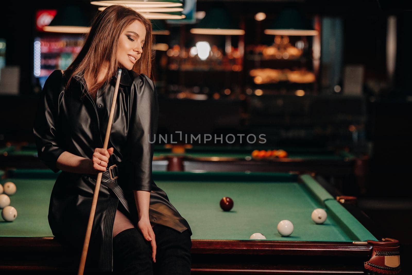 A girl with a cue in her hands sits on a table in a billiard club.Russian billiards by Lobachad