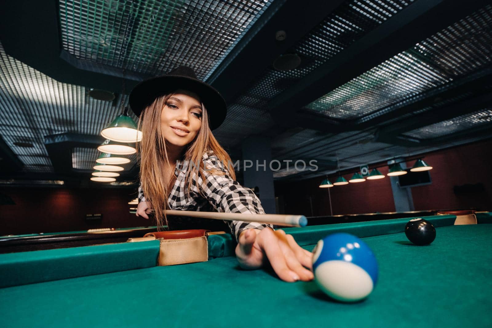 A girl in a hat in a billiard club with a cue in her hands hits a ball.Playing pool by Lobachad