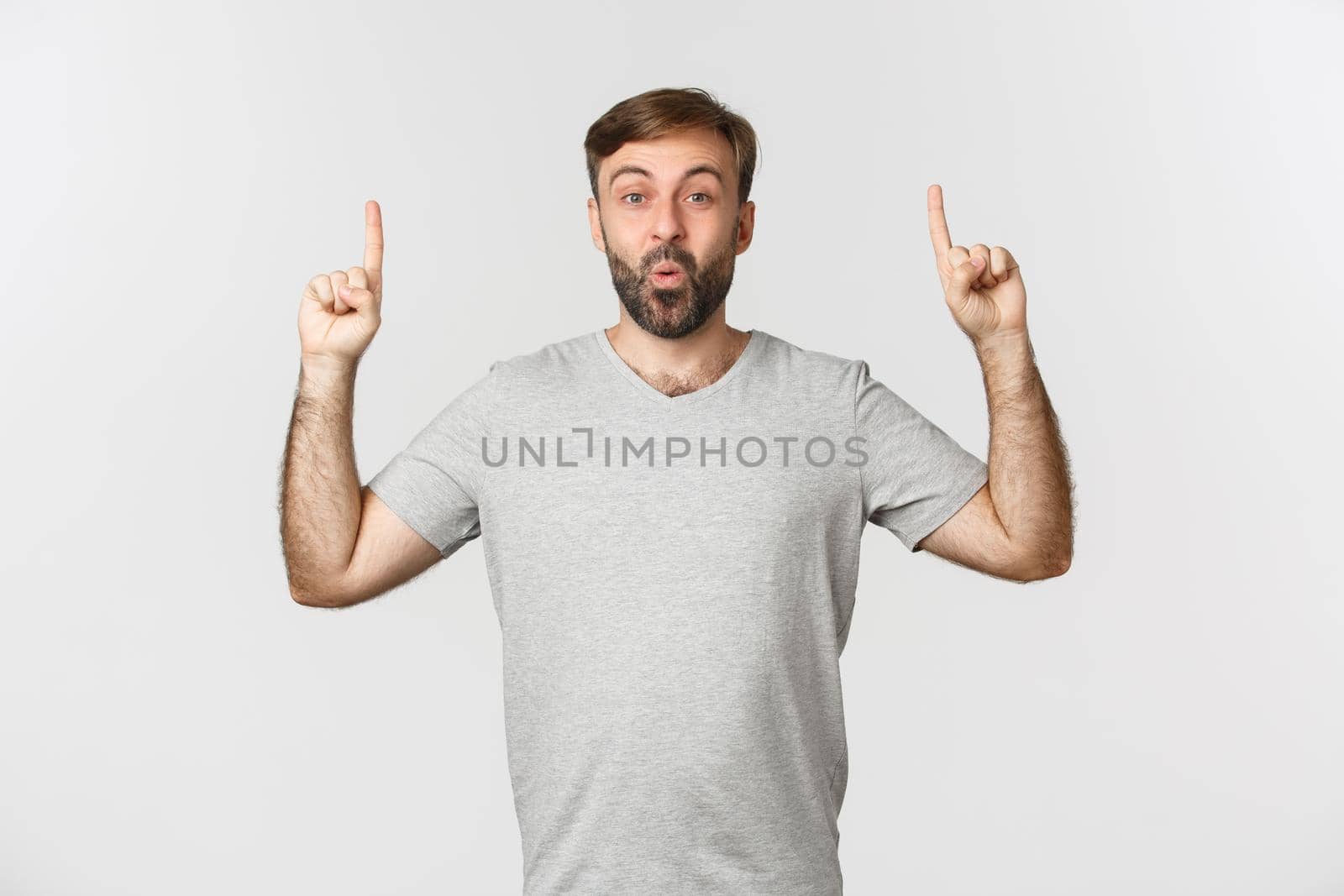 Image of excited handsome man in gray t-shirt, pointing fingers up and saying wow, standing over white background.