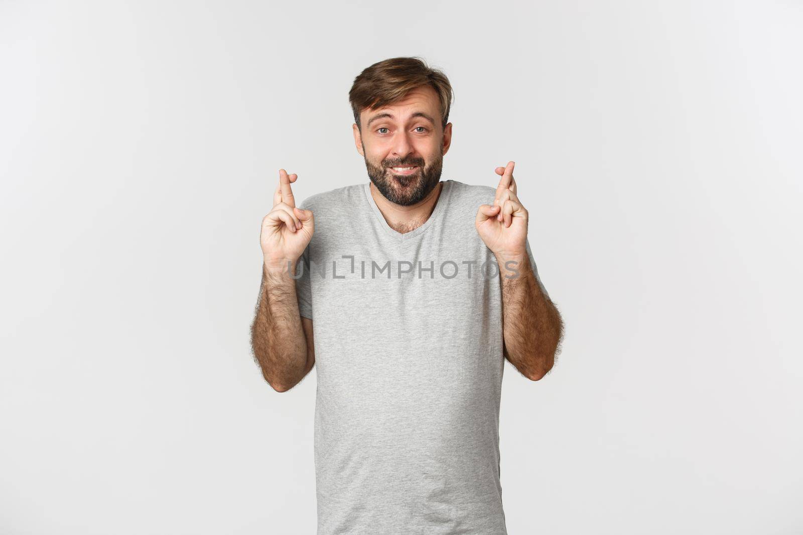 Portrait of hopeful smiling man with beard, wearing gray t-shirt, crossing fingers for good luck and looking at camera, making wish, standing over white background.