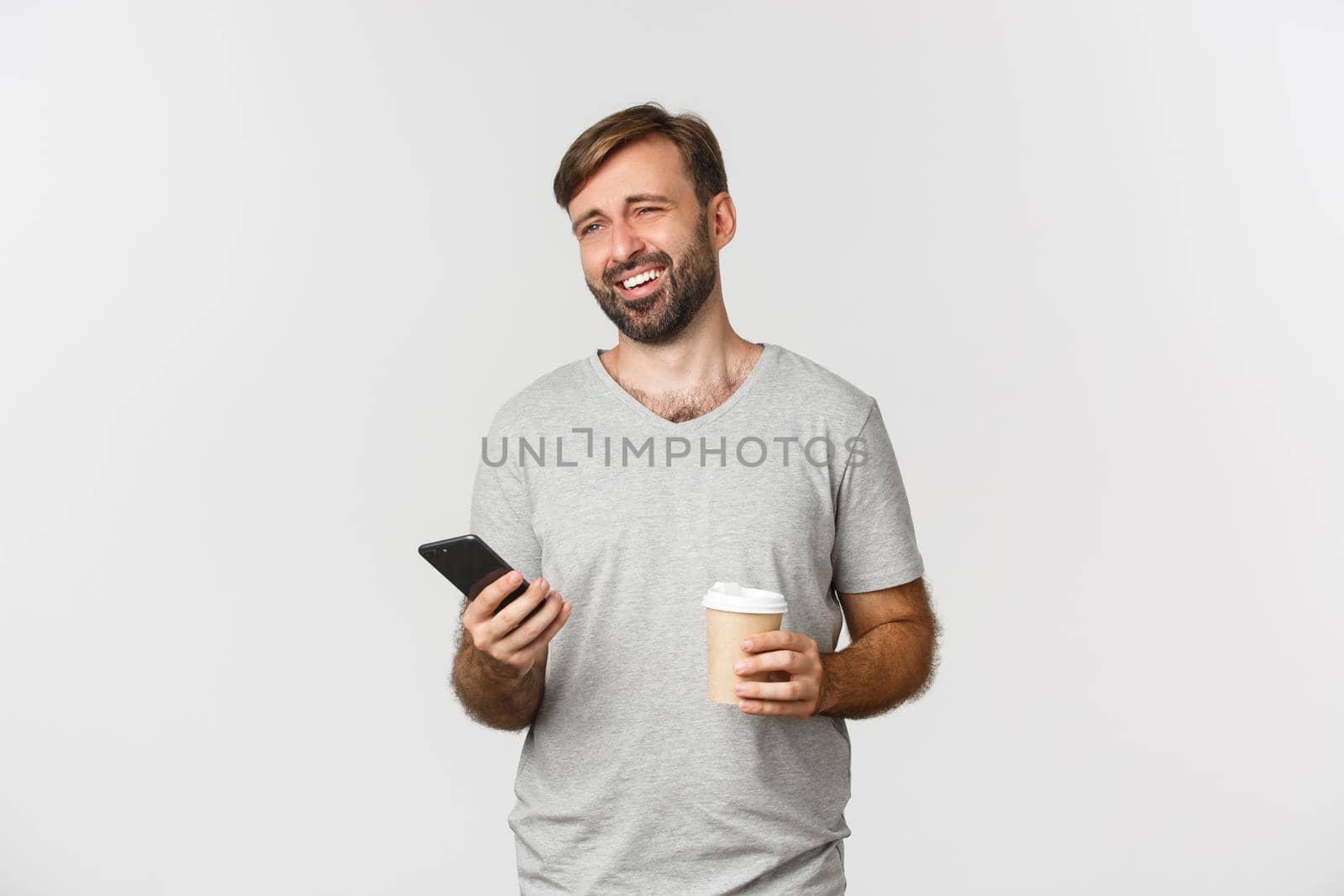 Image of modern caucasian man with beard, drinking coffee and casually using mobile phone, laughing as if talking to someone, standing over white background.