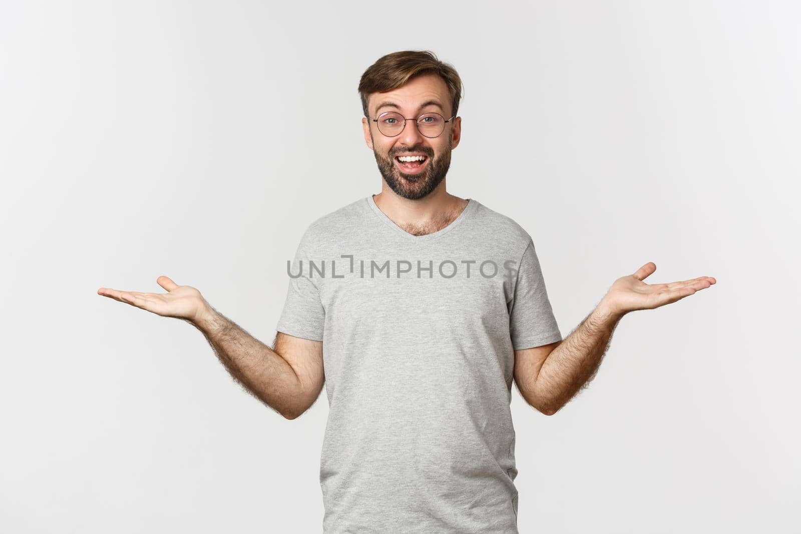 Handsome man with beard, wearing gray t-shirt and glasses, spread hands sideways and holding two things, demonstrate products, standing over white background.