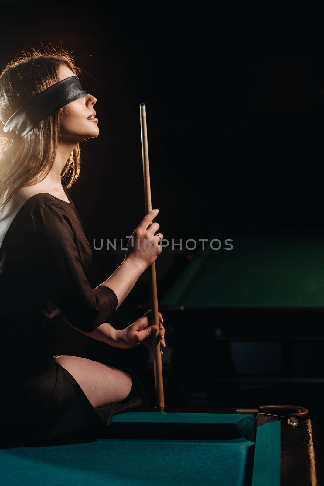 A girl with a blindfold and a cue in her hands is sitting on a table in a billiard club.Russian billiards.