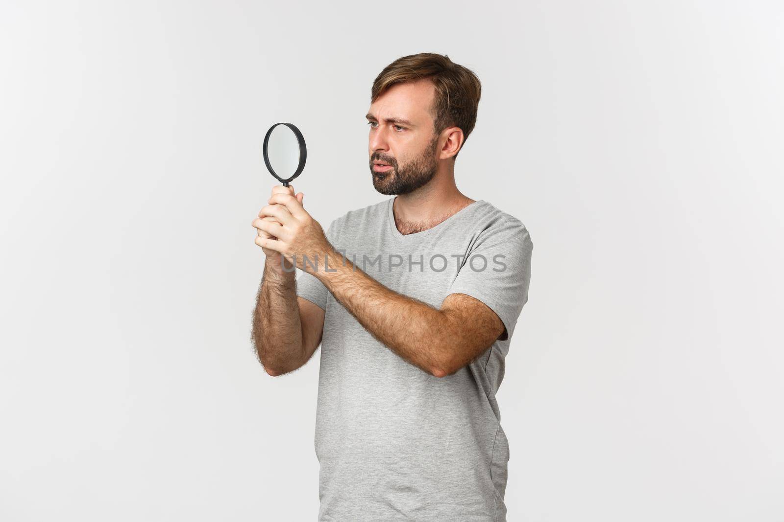 Image of handsome caucasian man in gray t-shirt, searching for something, looking through magnifying glass with curious face, standing over white background.