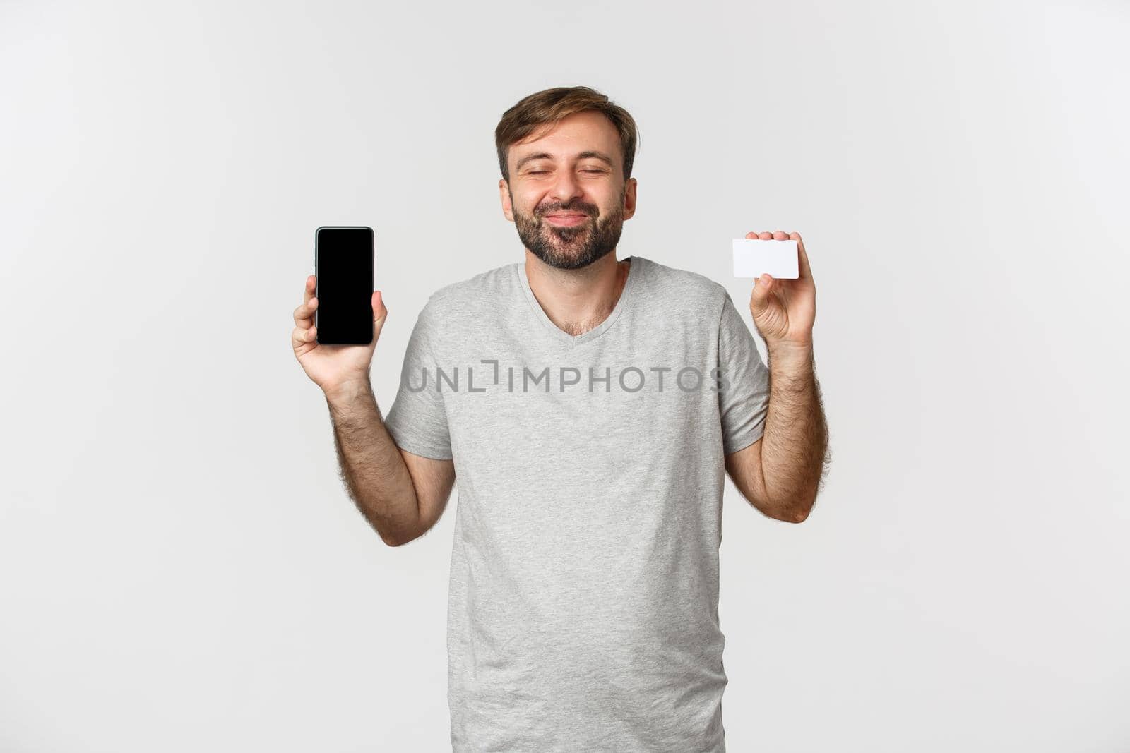Handsome young man shopping online, holding credit card and mobile phone, showing smartphone screen, standing over white background.