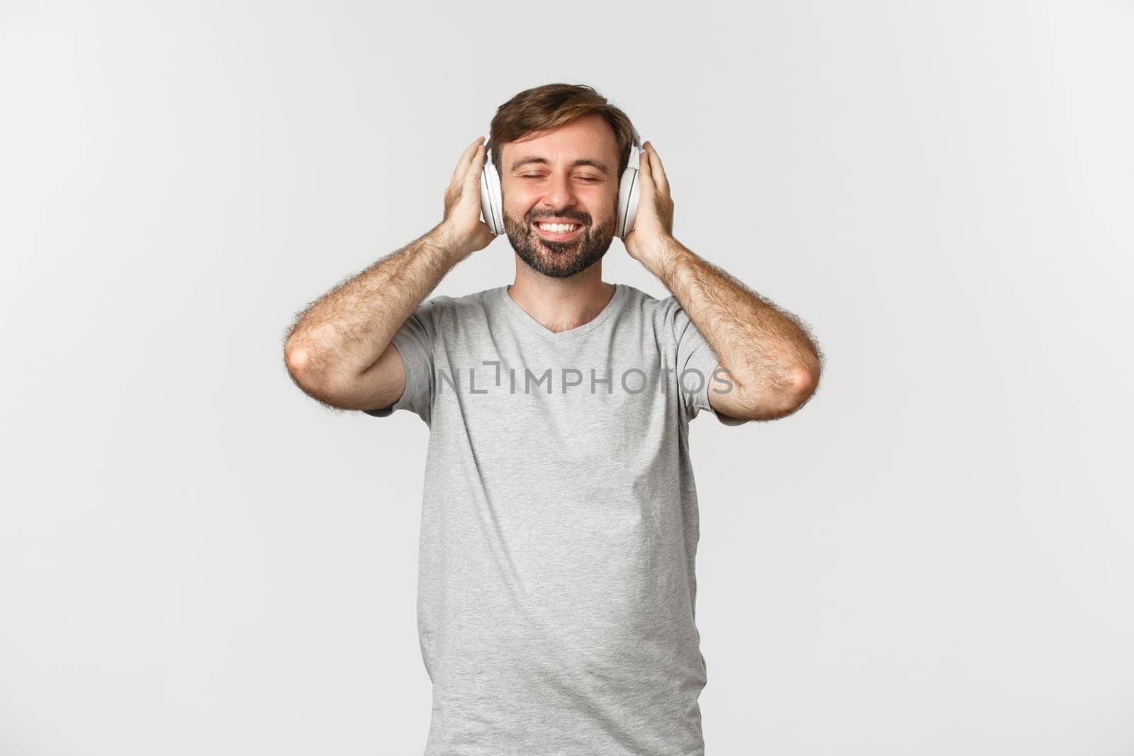 Image of handsome man in casual outfit, listening music in wireless headphones, smiling pleased, standing over white background by Benzoix