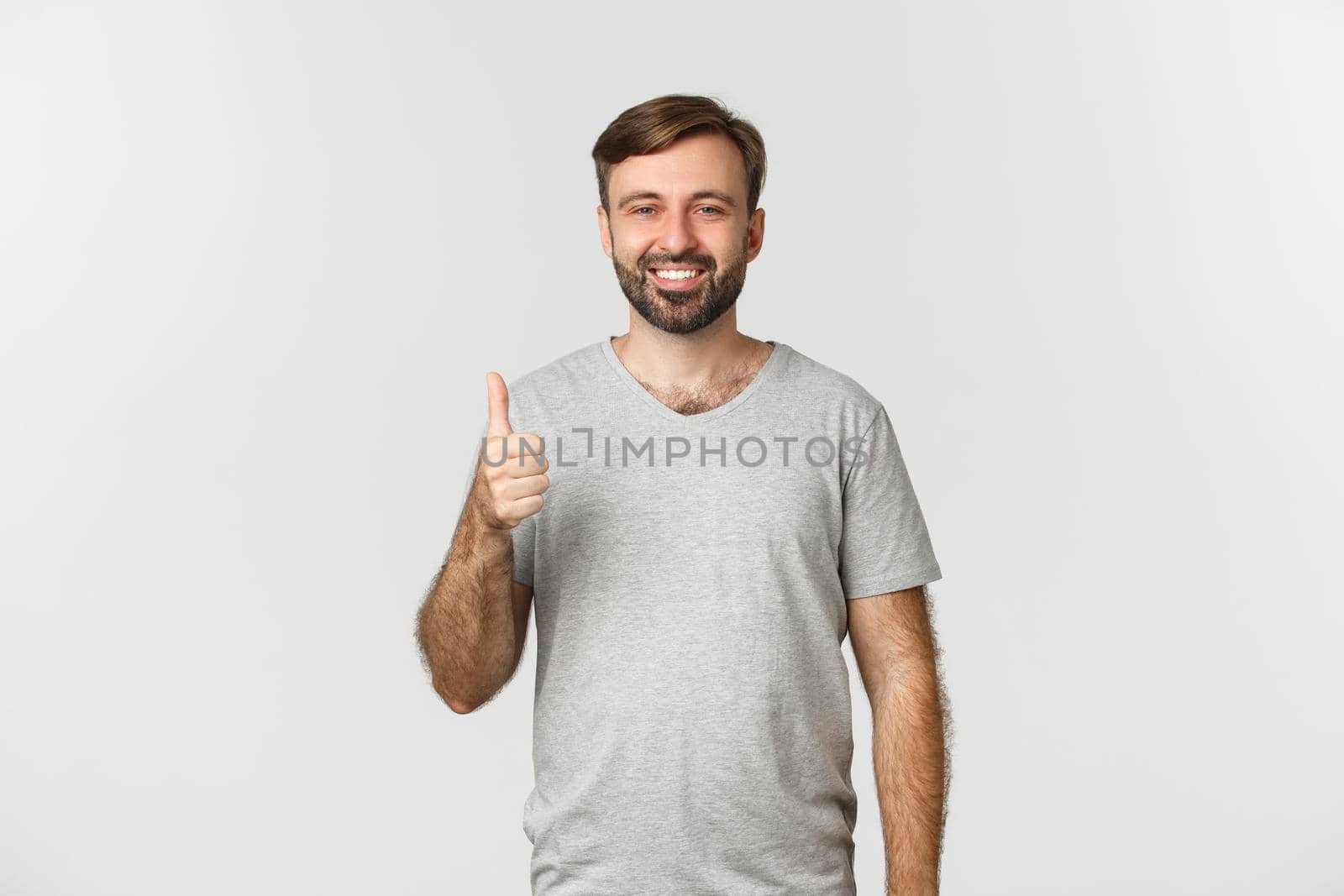 Image of satisfied smiling man in gray t-shirt, showing thumbs-up in approval, recommend something good, white background.