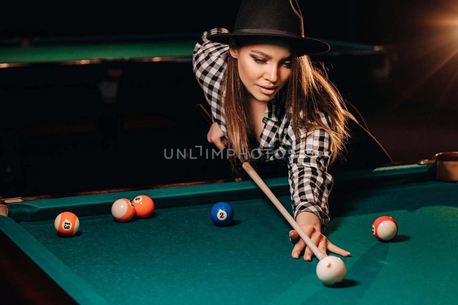 A girl in a hat in a billiard club with a cue in her hands hits a ball.Playing billiards.