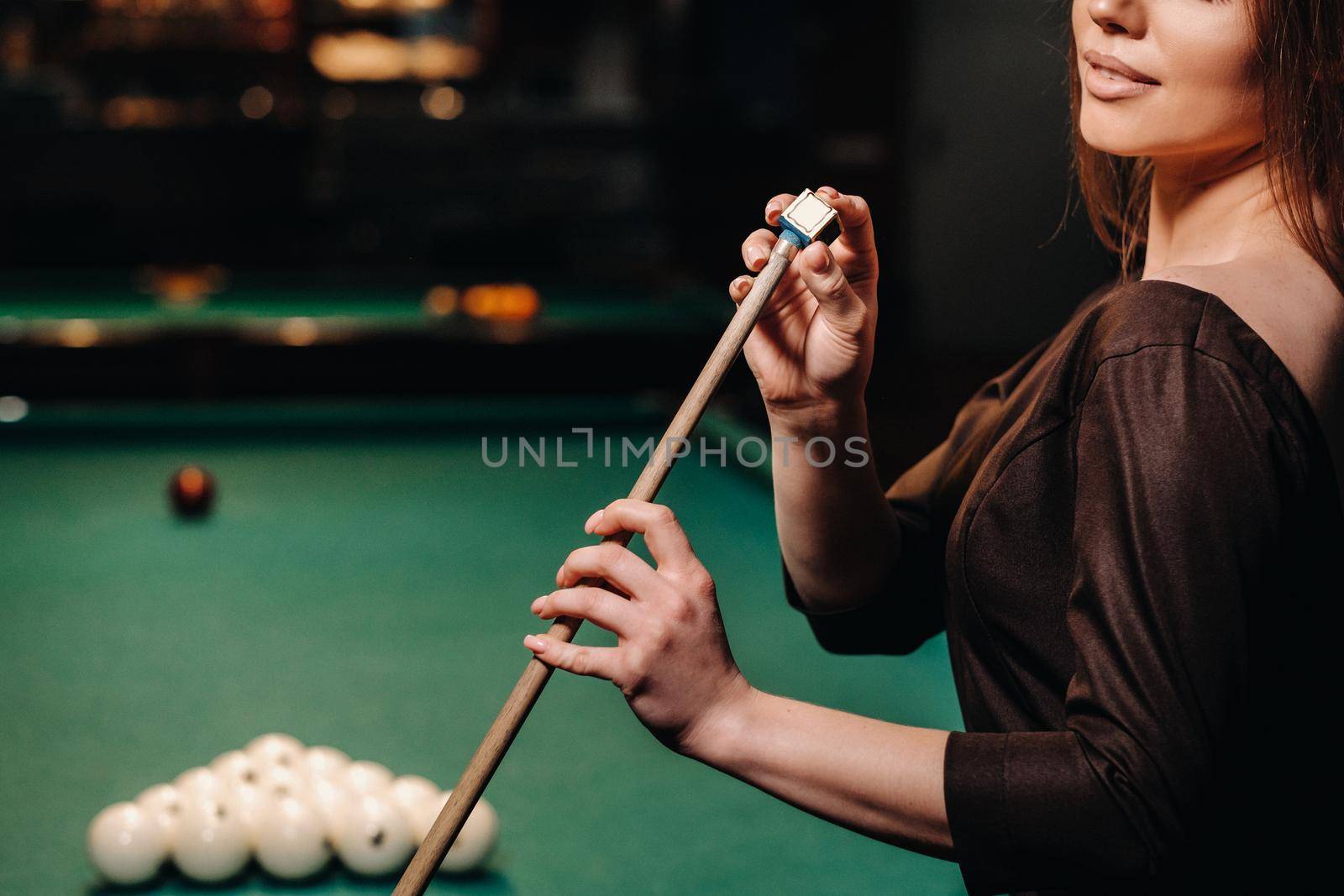 A girl in a dress stands with a cue in her hands and cleans it with chalk in a billiard club with balls in her hands.Playing billiards by Lobachad