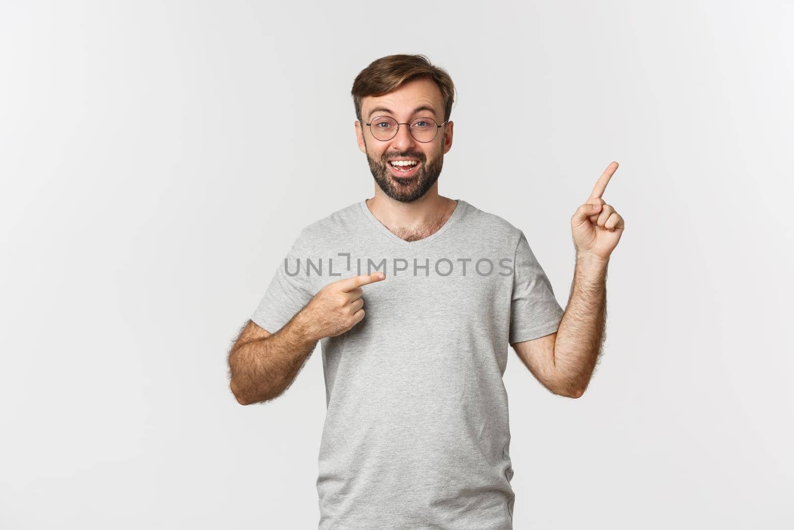 Surprised bearded man smiling, pointing fingers at upper right corner, showing logo, standing over white background.