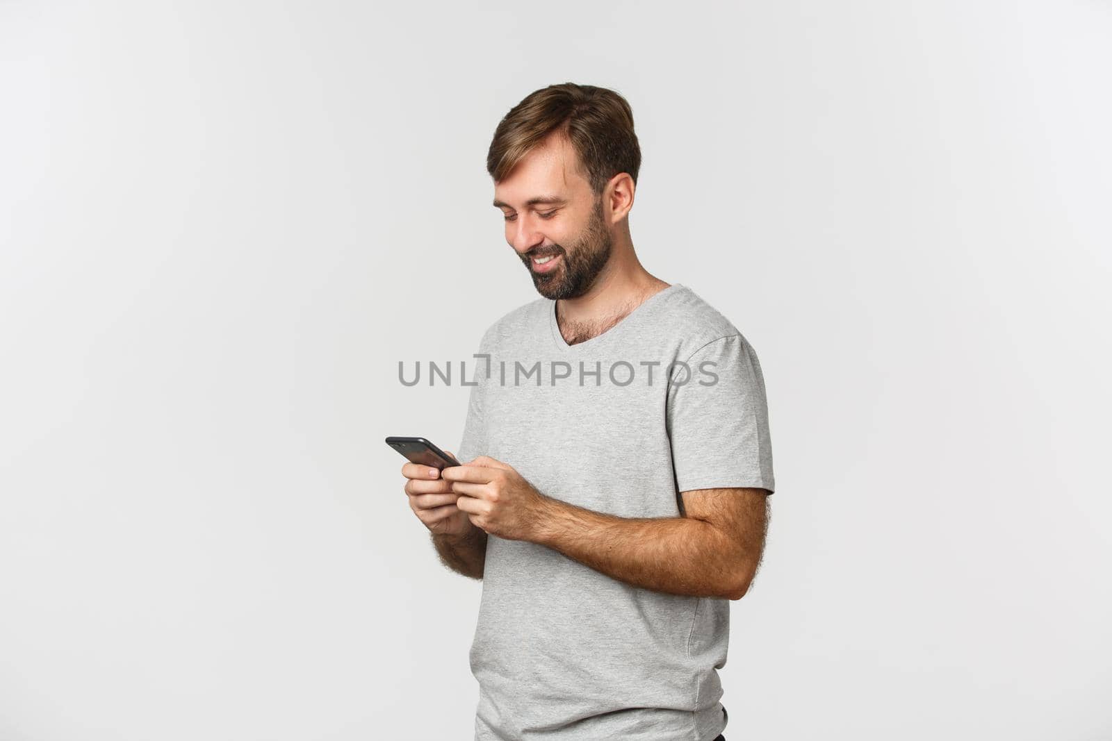 Portrait of modern bearded man in gray t-shirt, messaging and looking at smartphone happy, standing over white background by Benzoix
