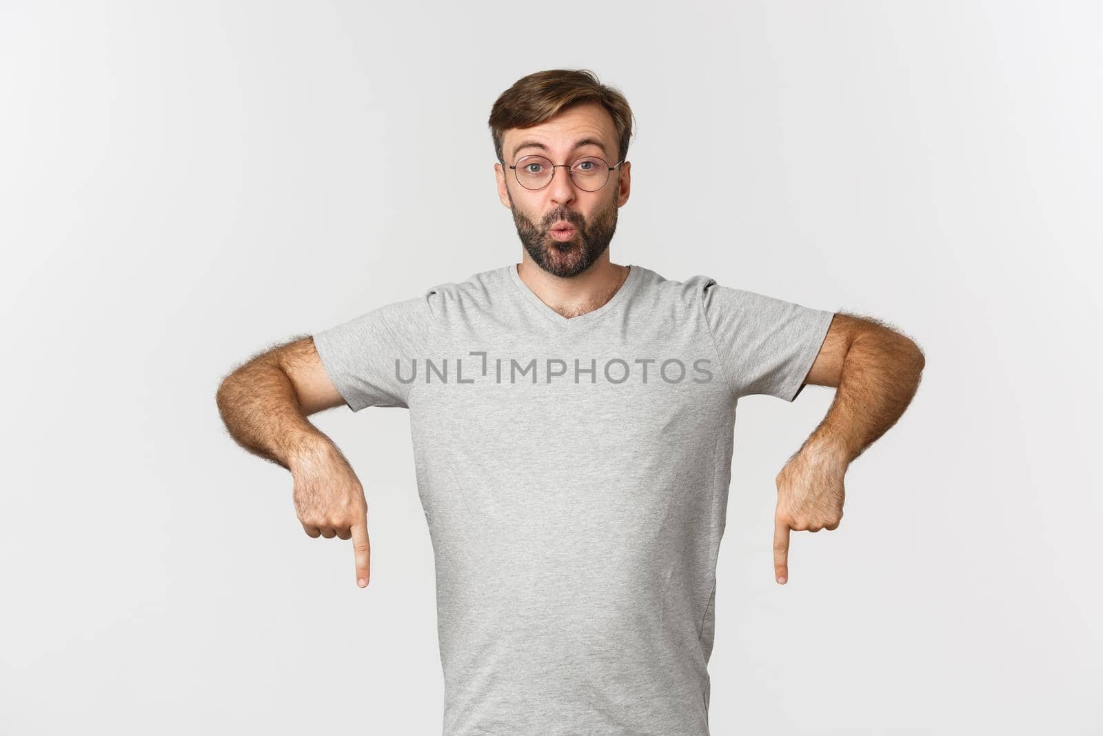 Surprised and excited bearded man, pointing fingers down, showing logo, wearing gray t-shirt, wearing gray t-shirt, standing over white background by Benzoix