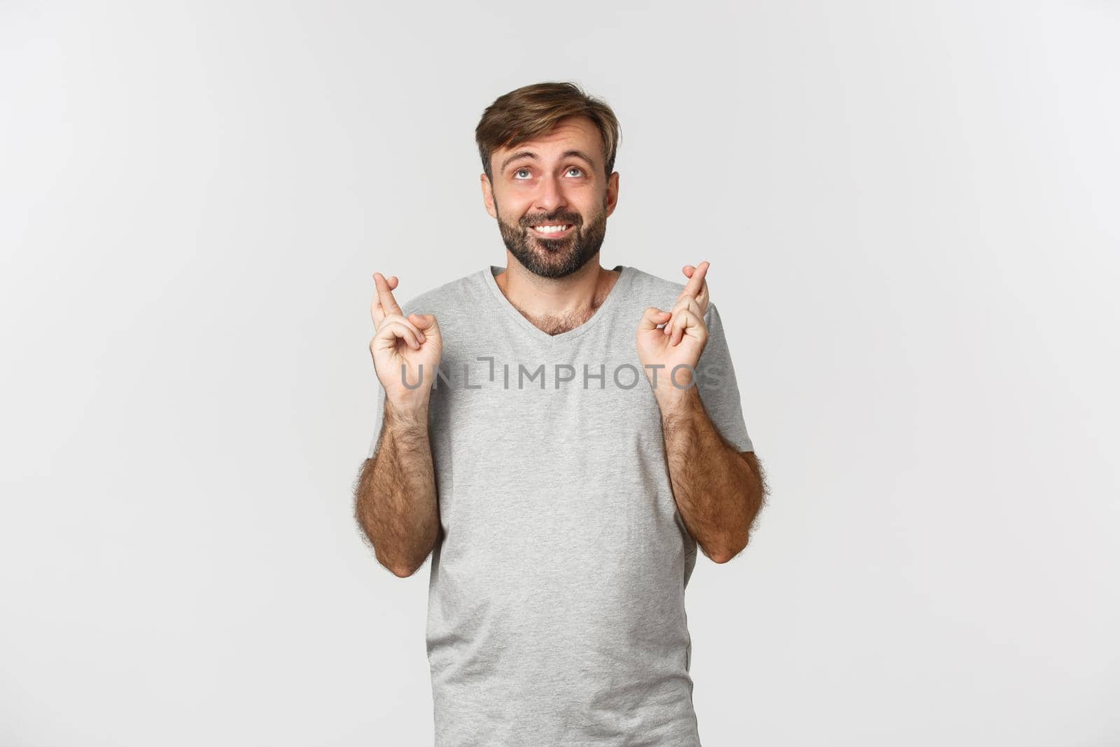 Hopeful smiling man in gray t-shirt, looking up and making wish, crossing fingers for good luck, standing over white background.