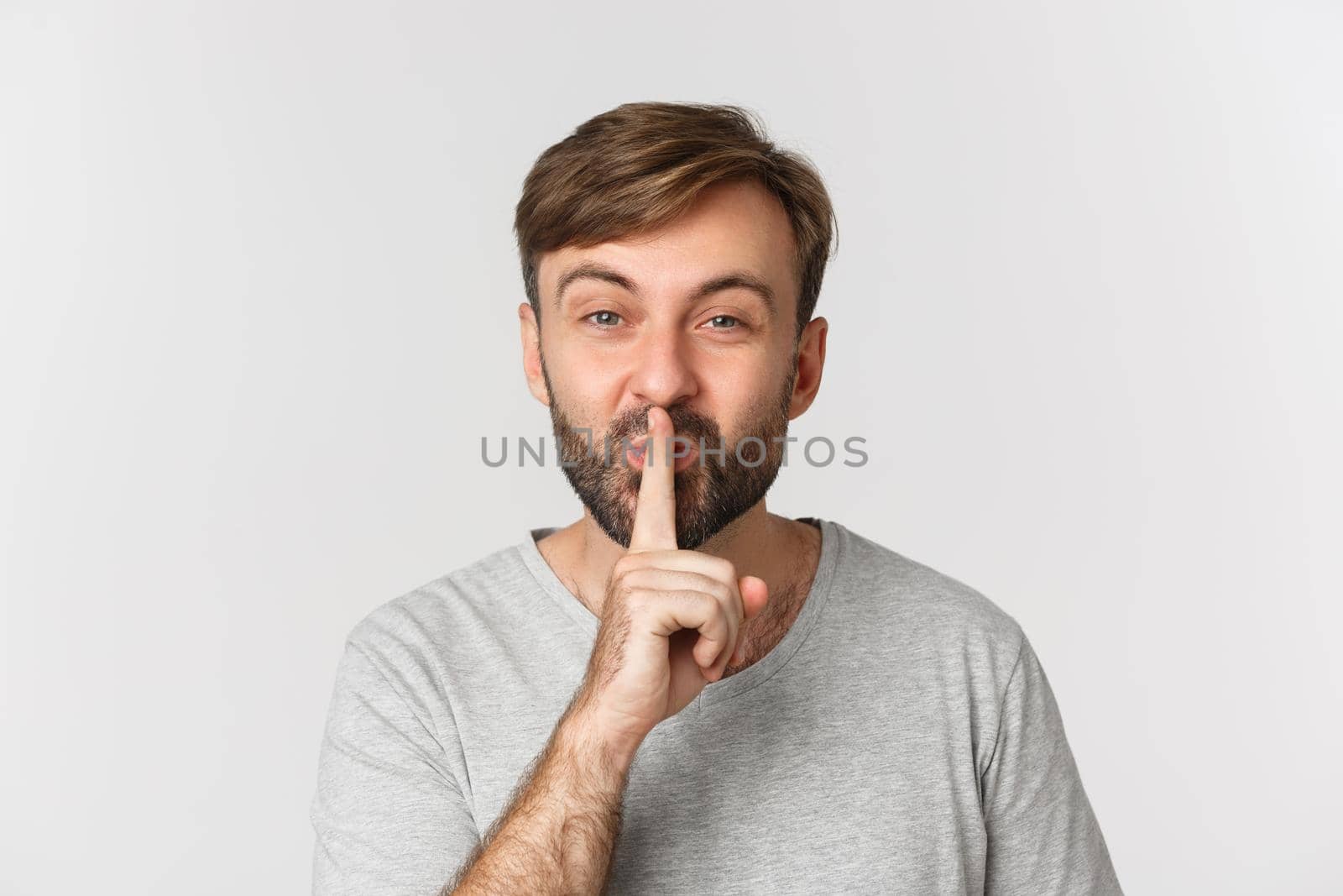 Close-up of smiling man in gray t-shirt, hushing, telling a secret, standing over white background by Benzoix