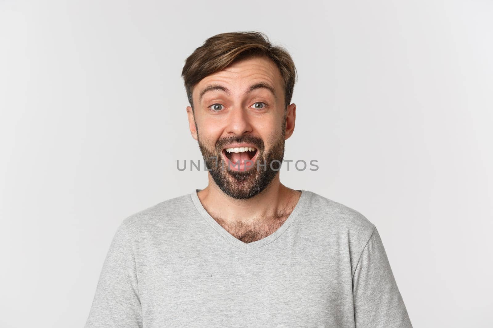 Close-up of handsome adult man with beard, wearing gray t-shirt, smiling surprised, standing over white background.