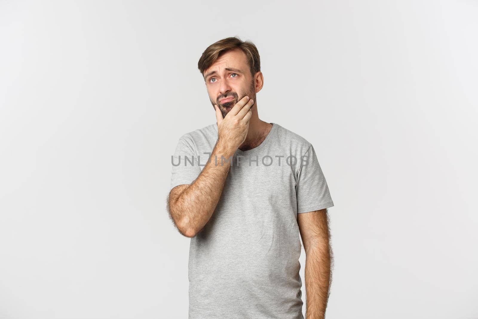 Troubled sad guy touching beard, looking at upper left corner thoughtful, standing indecisive over white background.