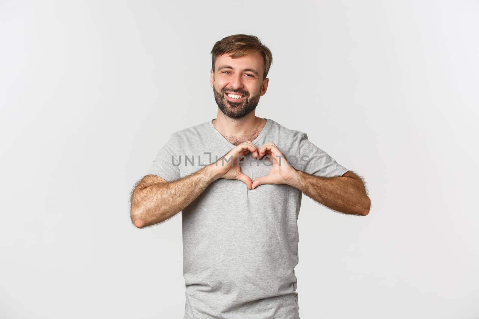 Portrait of handsome smiling man with beard, showing heart sign, love someone, standing over white background.