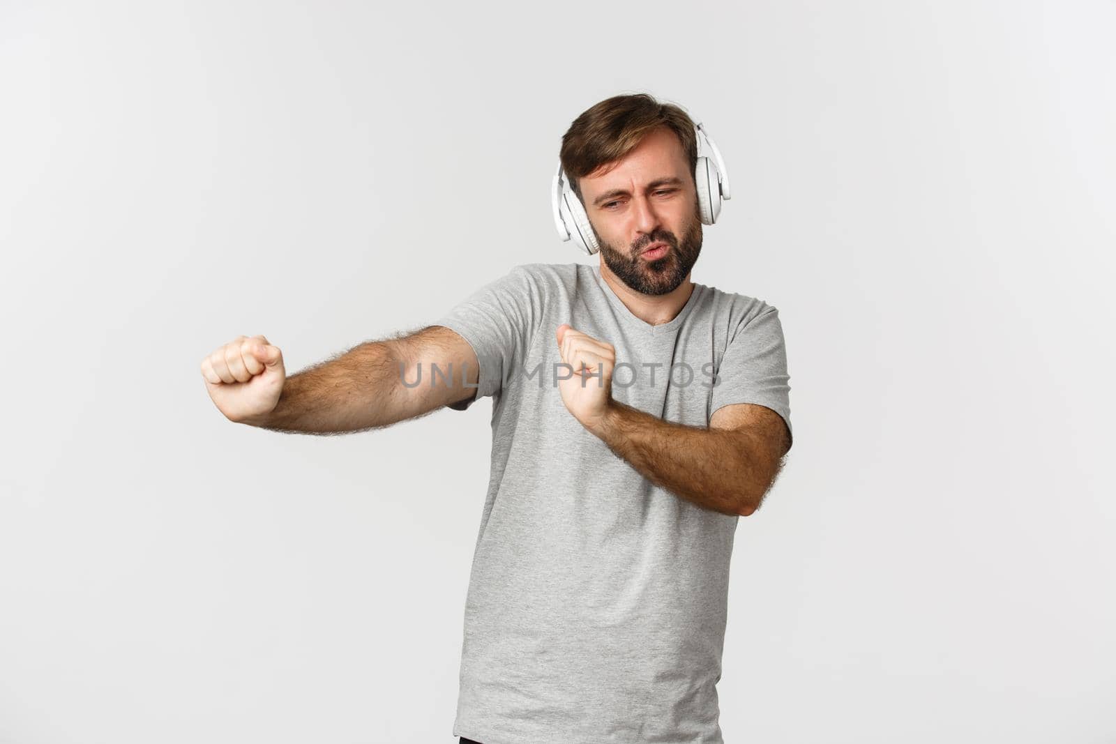 Portrait of happy handsome guy in gray t-shirt, listening music in wireless headphones and dancing, standing over white background.