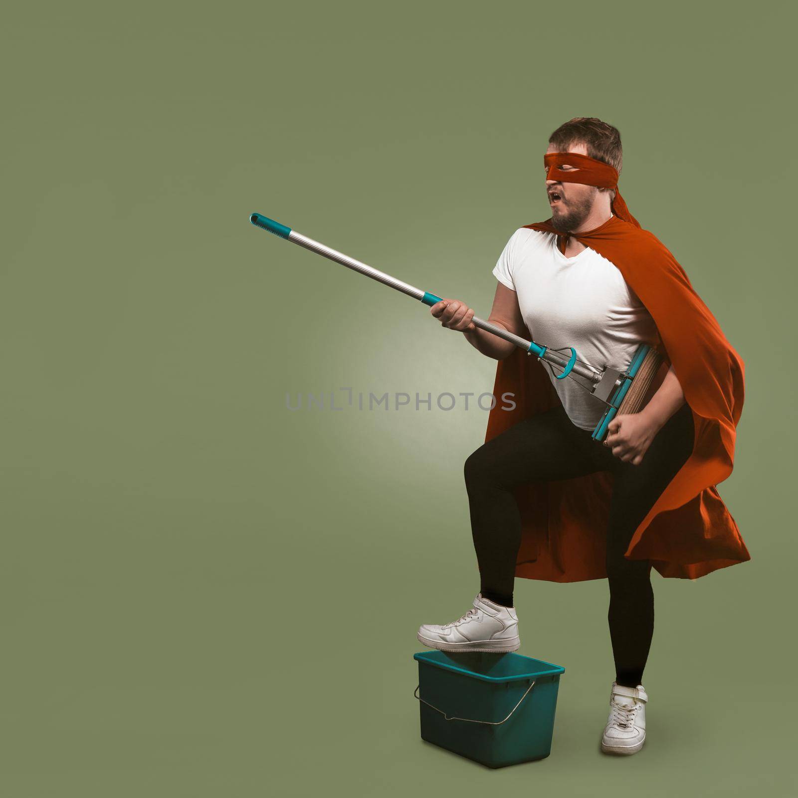 Superhero has fun with cleaning equipment. Man in red cape holds mop and presents himself as musician with guitar. Isolated on chive background. Template with copy space at left side.