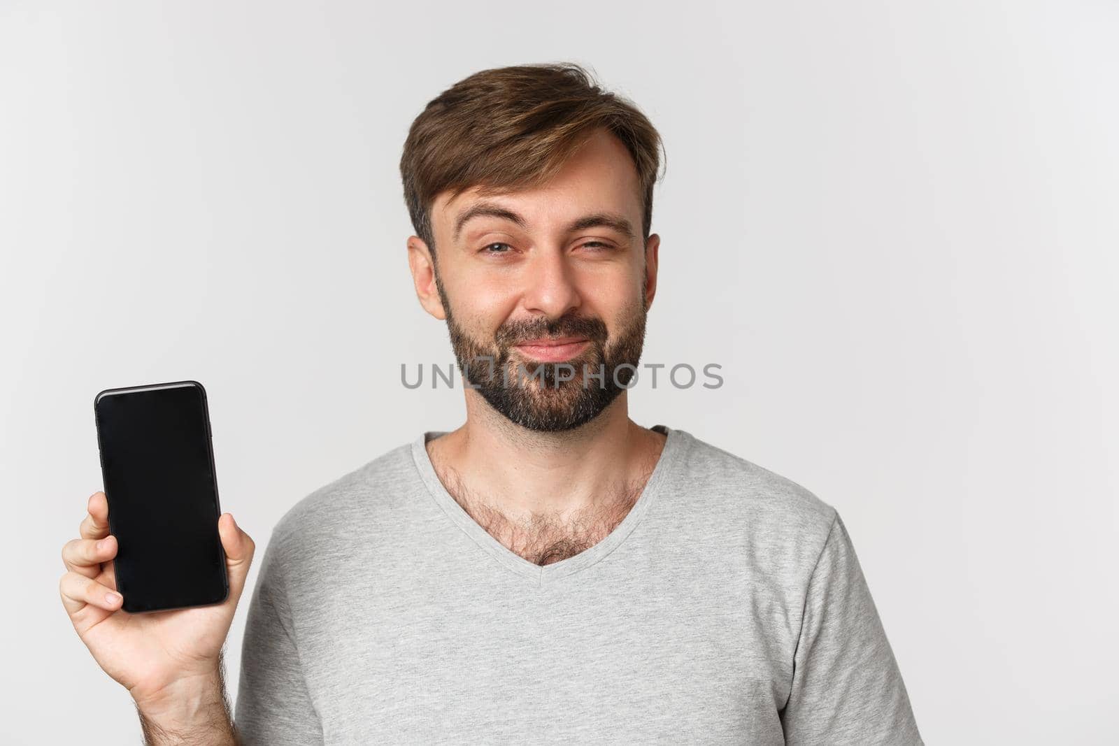 Close-up of pleased smiling man with beard, looking delighted and showing mobile phone screen, standing over white background.